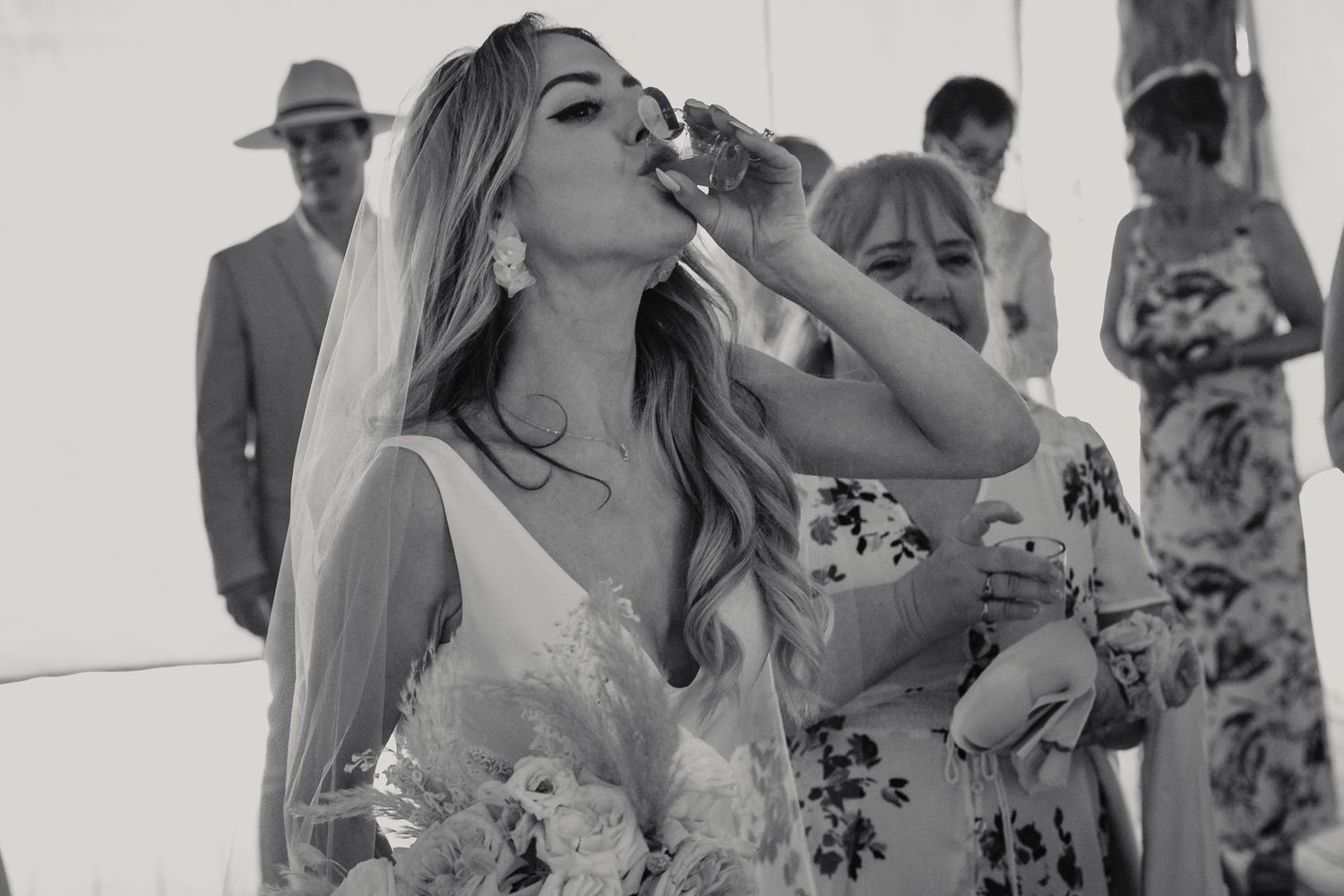 Bride in white gown and veil drinks from a glass while holding bouquet at outdoor wedding ceremony.