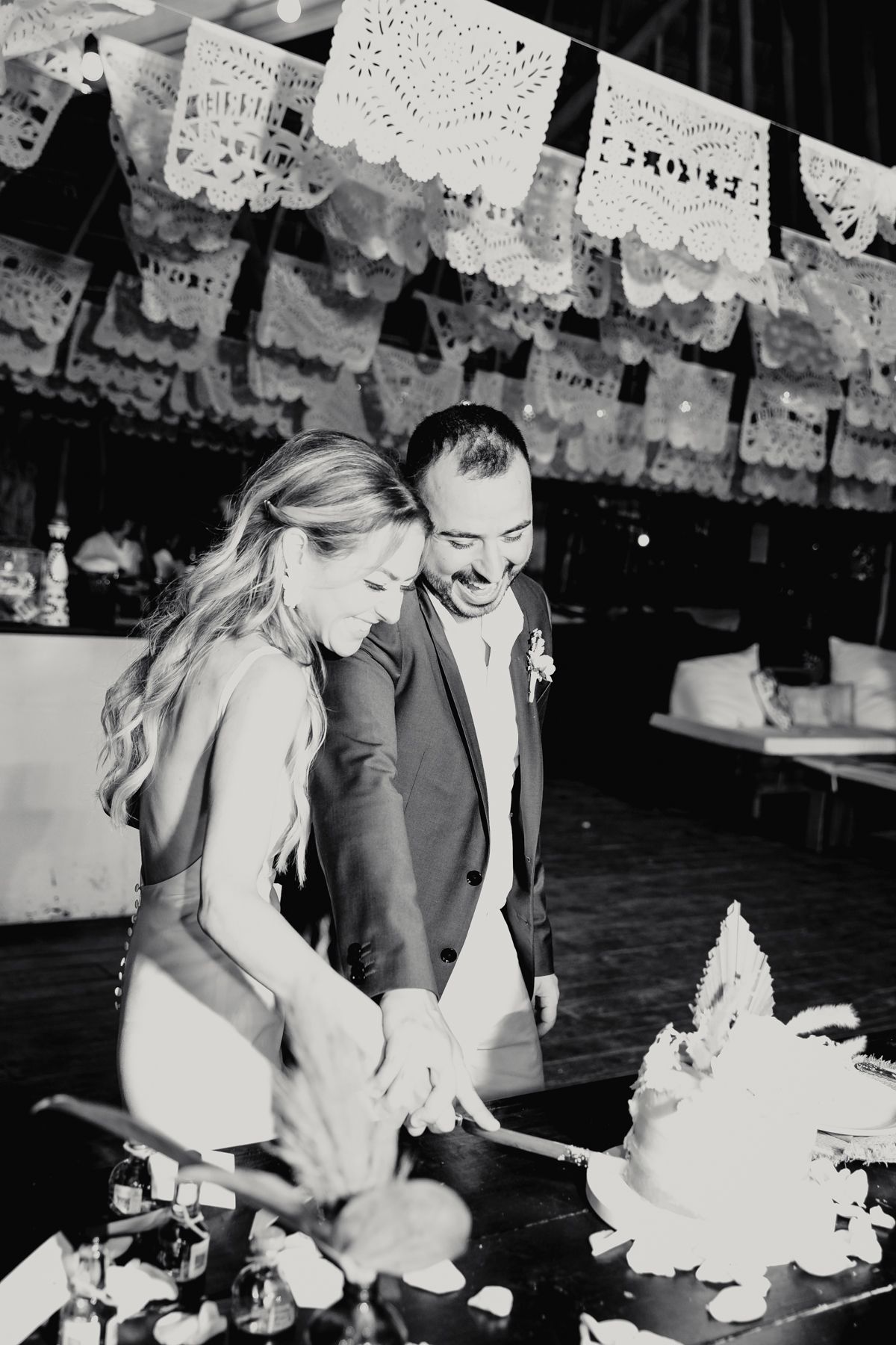 Bride and groom cut wedding cake together under decorative papel picado banners at festive reception in black and white.