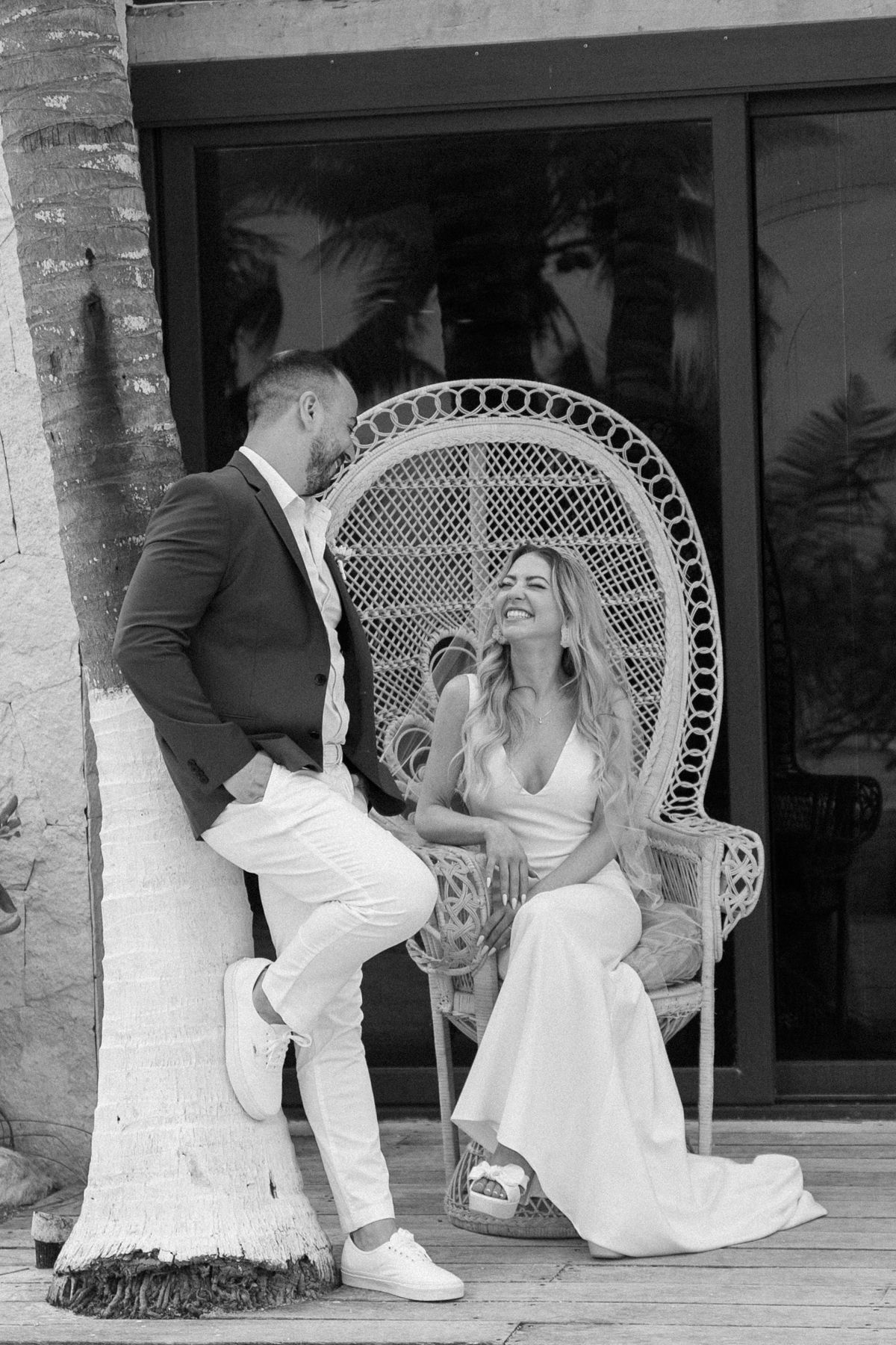 Bride and groom share a joyful moment beside a wicker peacock chair and palm tree in a black and white wedding photo.