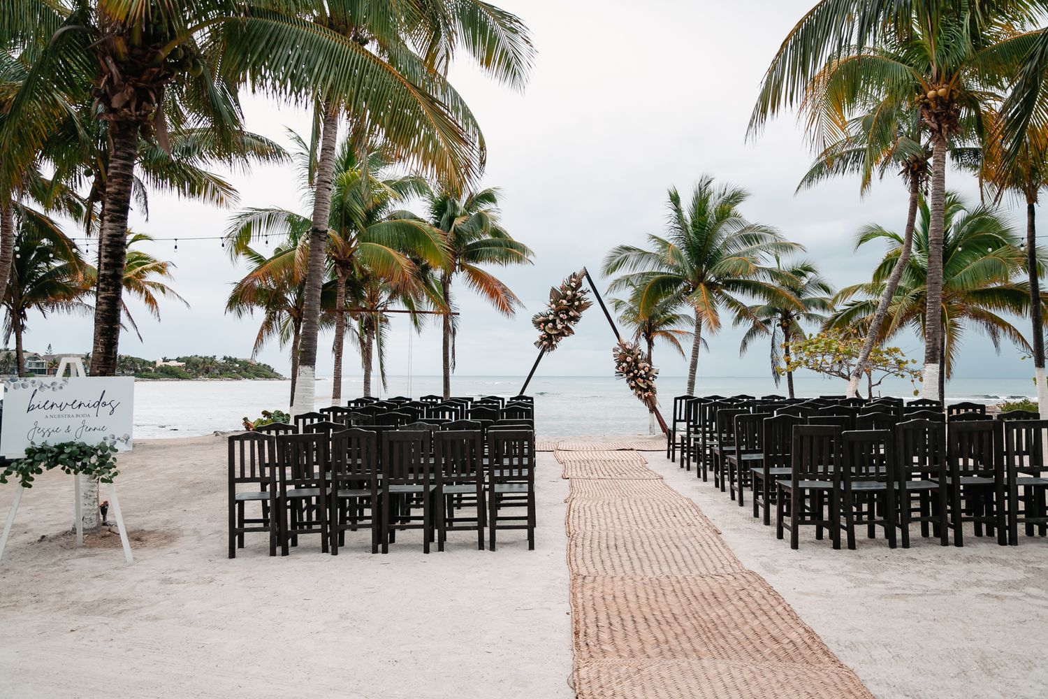 Outdoor beach wedding ceremony setup with black chairs, brick aisle, floral arch, and palm trees by the ocean.