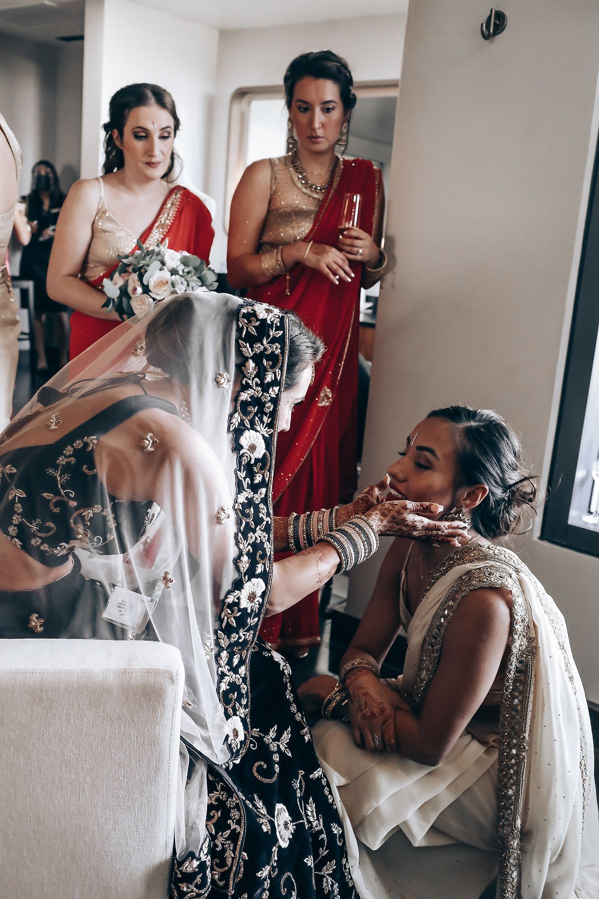 A group of women in traditional Indian wedding attire gather in a bright room during wedding preparations.