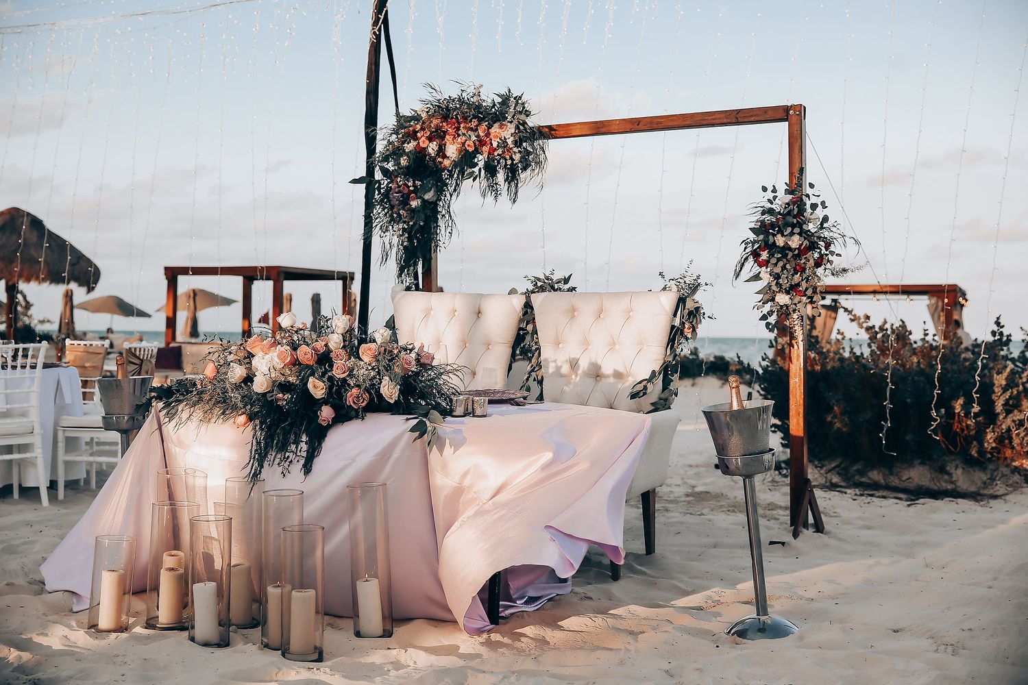 Outdoor wedding venue setup with wooden arches, white fabric draping, and floral decorations at sunset.