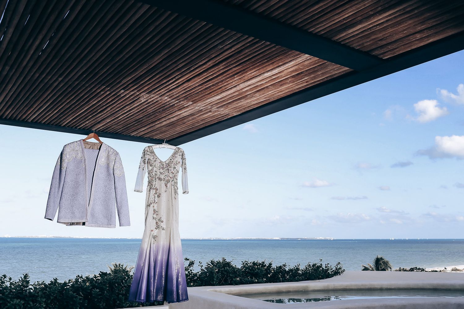 Wedding dress and suit hanging from wooden pergola overlooking ocean view with blue sky and clouds.
