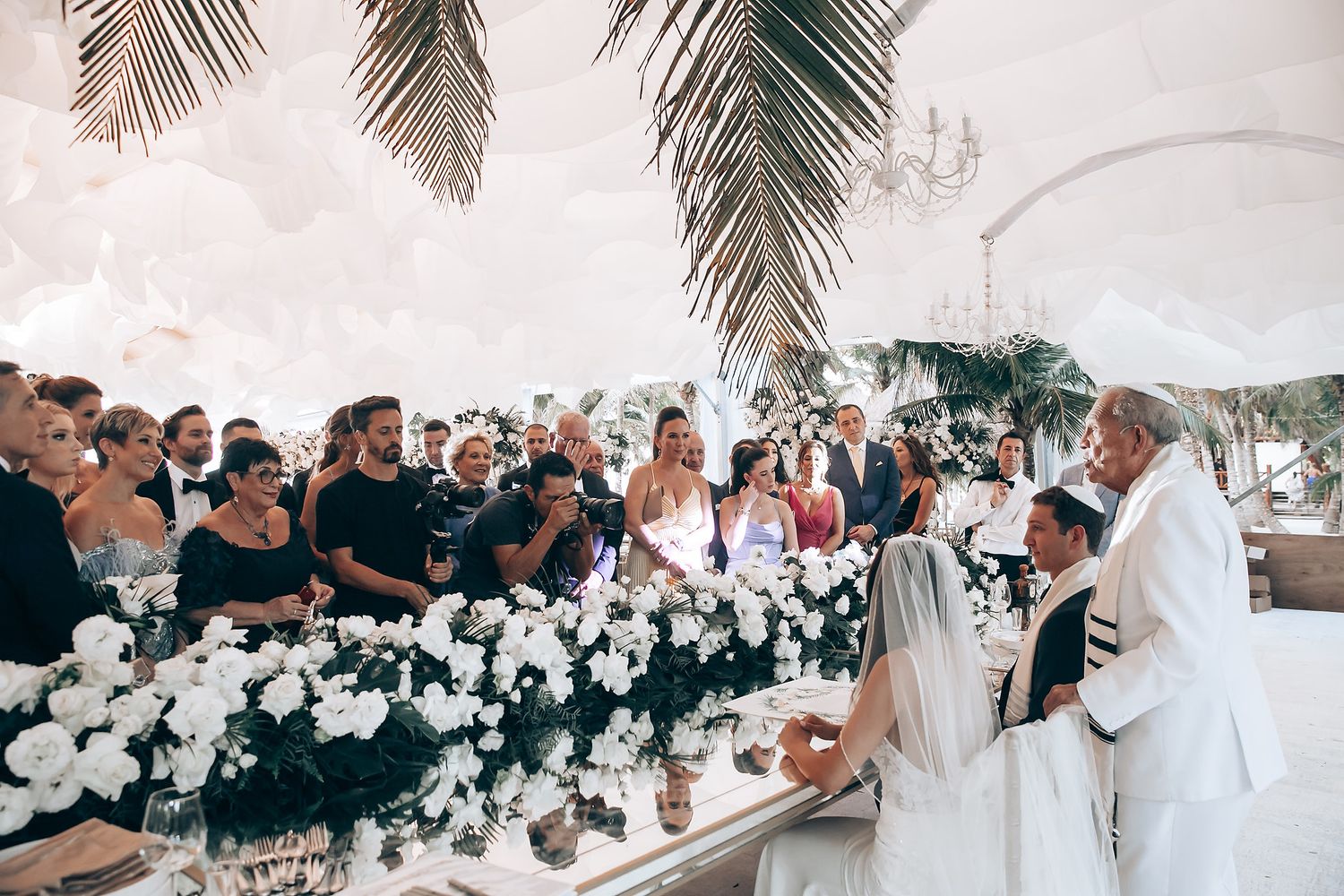 Wedding reception guests seated at long tables under white tents decorated with floral arrangements and string lights.