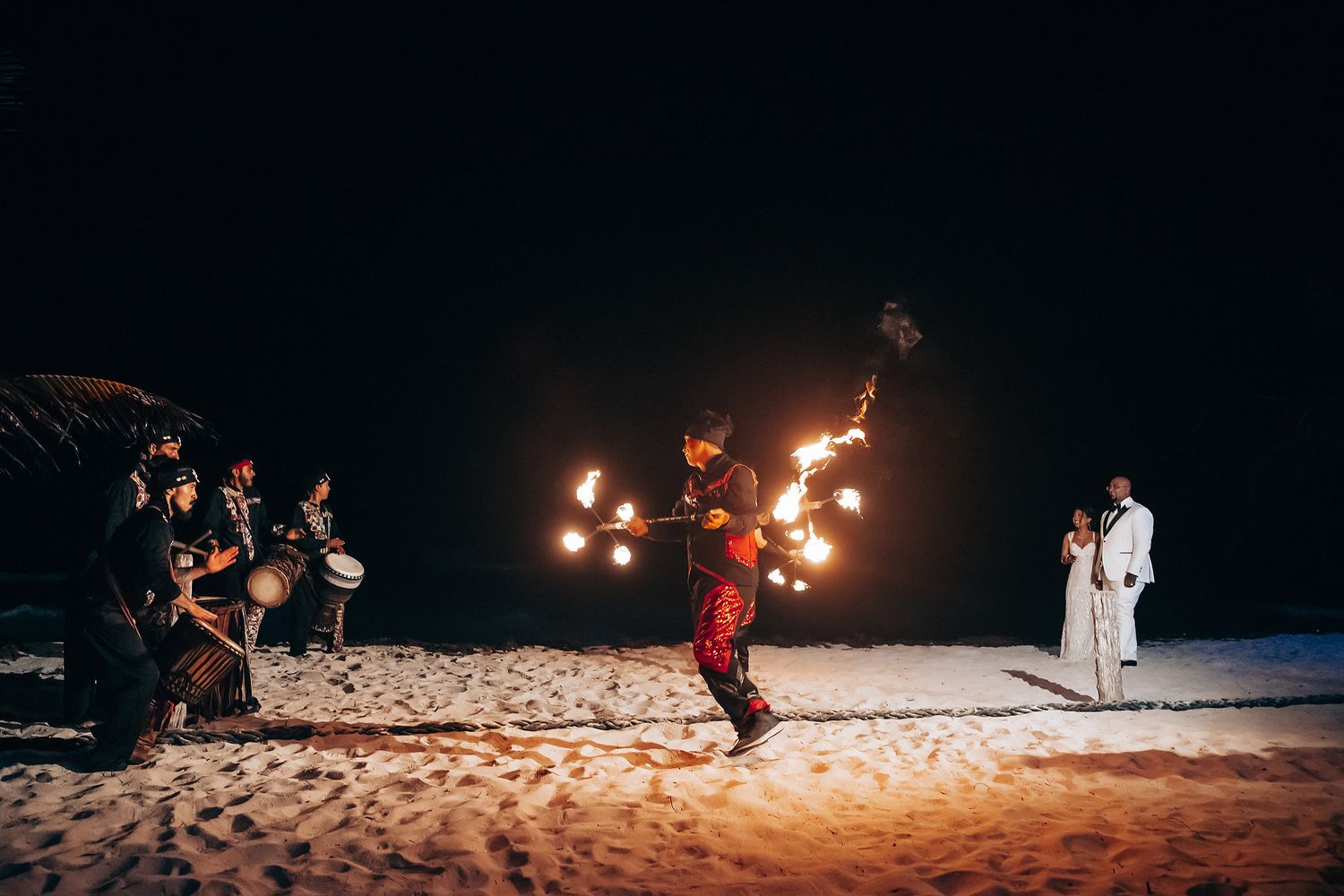 Fire performers entertain guests on a beach at night with glowing flames.