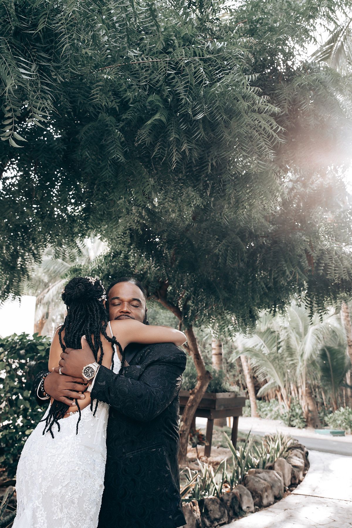 Two people share an intimate embrace under lush green trees during a wedding ceremony at an outdoor venue.