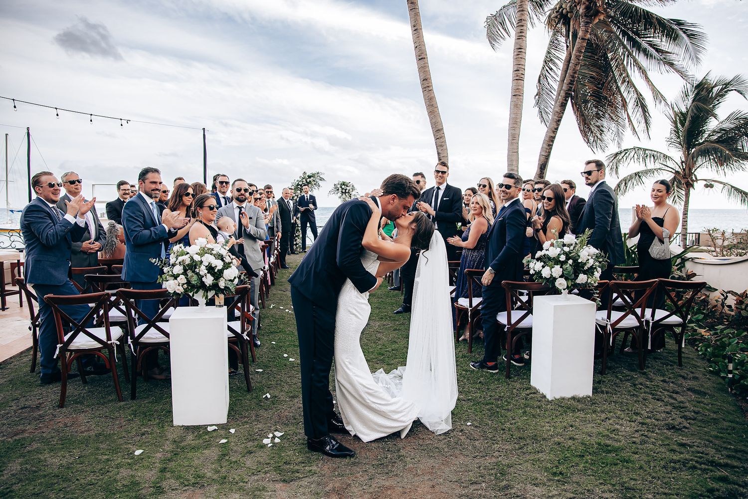 Wedding reception setup on beach with palm trees and string lights at sunset.
