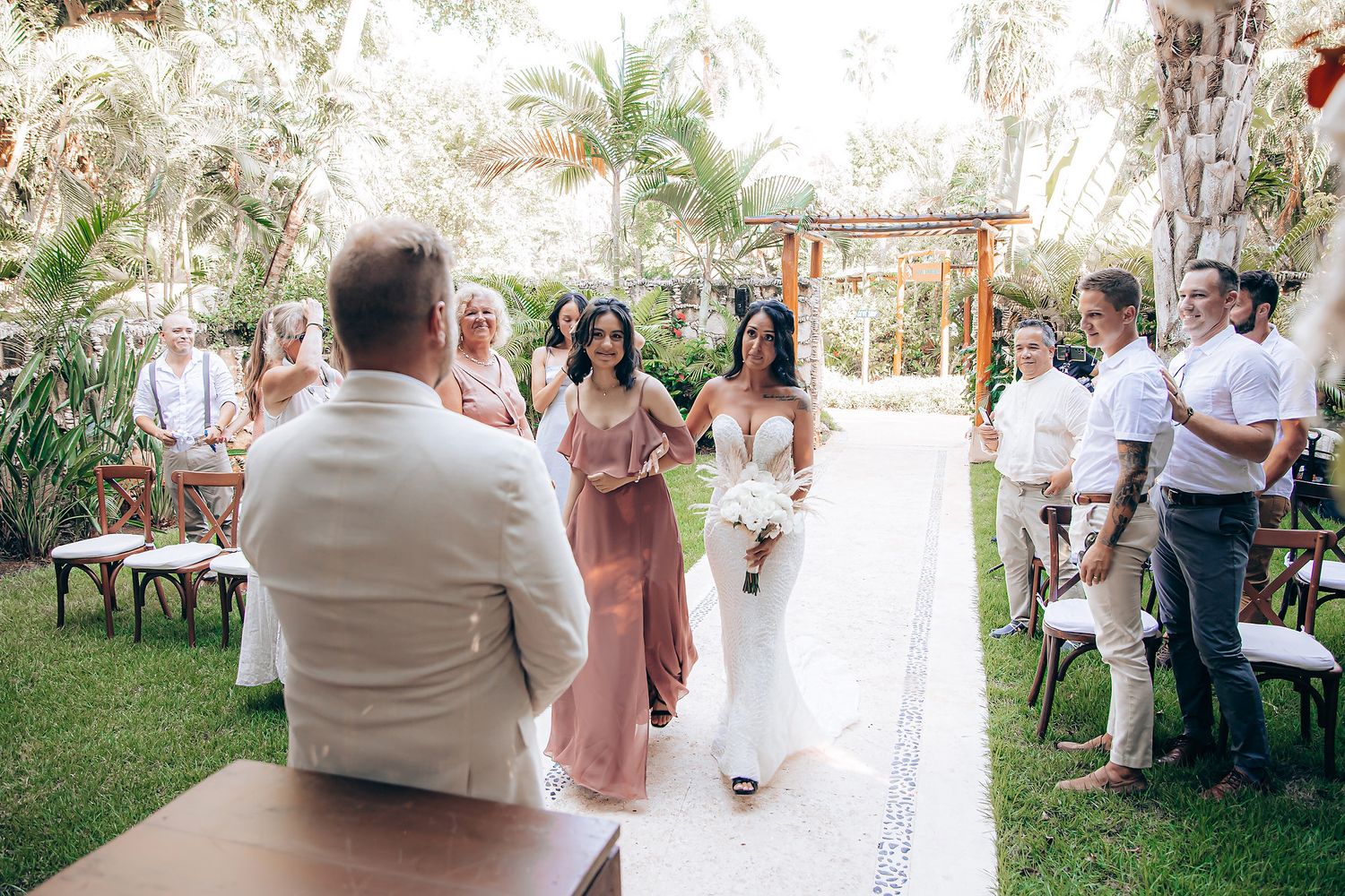 Outdoor wedding ceremony in a tropical garden setting with guests seated on wooden chairs.