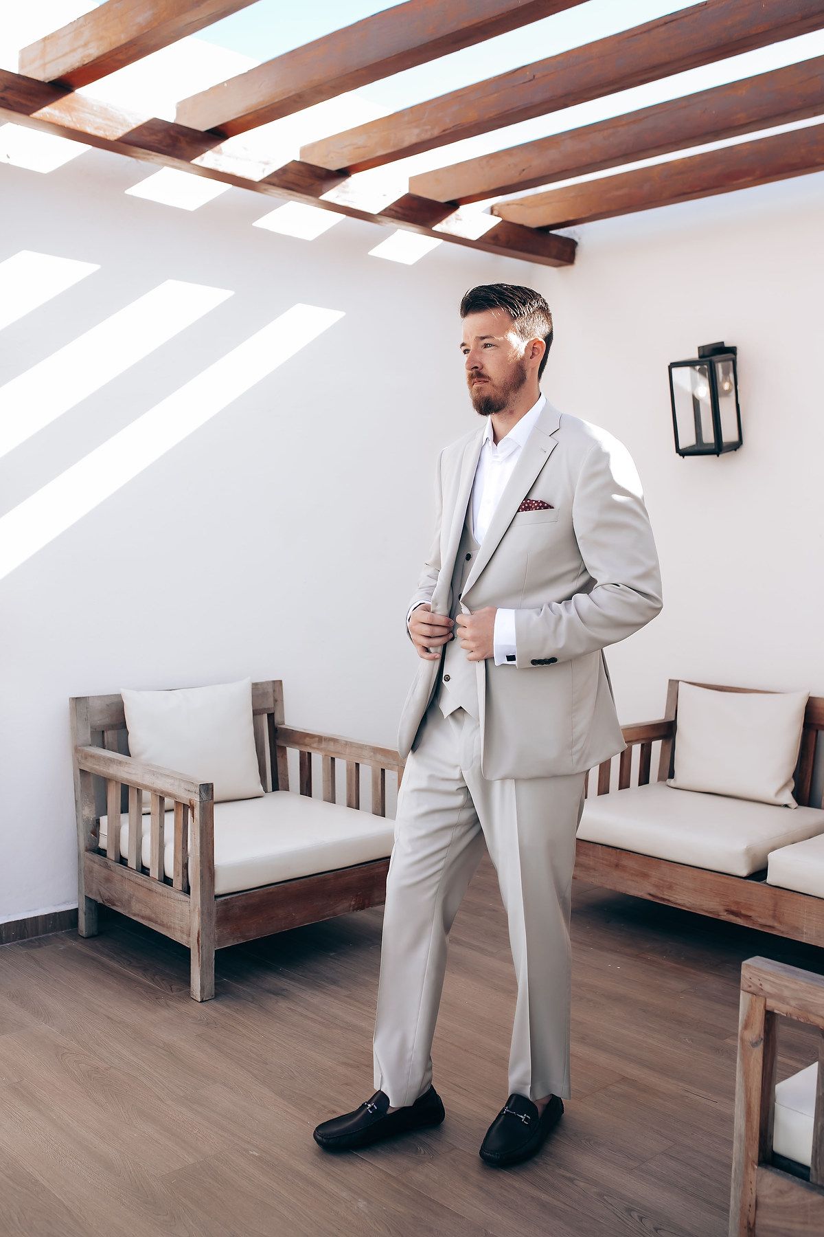 Man in beige suit stands in elegant room with wooden beams and bench seating.