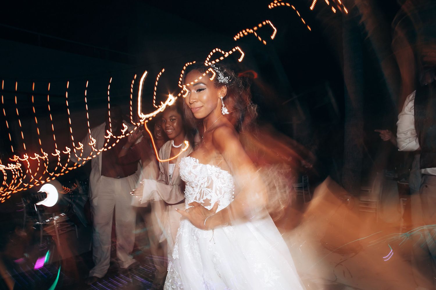 Wedding guests dance and celebrate on a dimly lit dance floor decorated with string lights above.