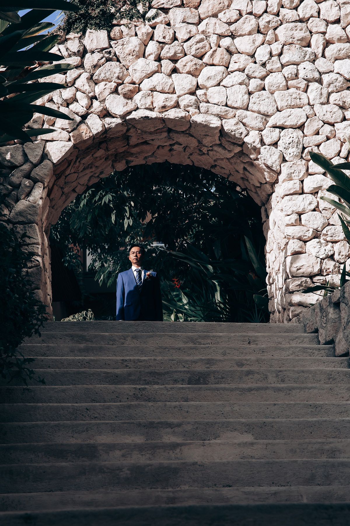 A figure stands in a stone archway on steps surrounded by tropical foliage at a scenic outdoor location.