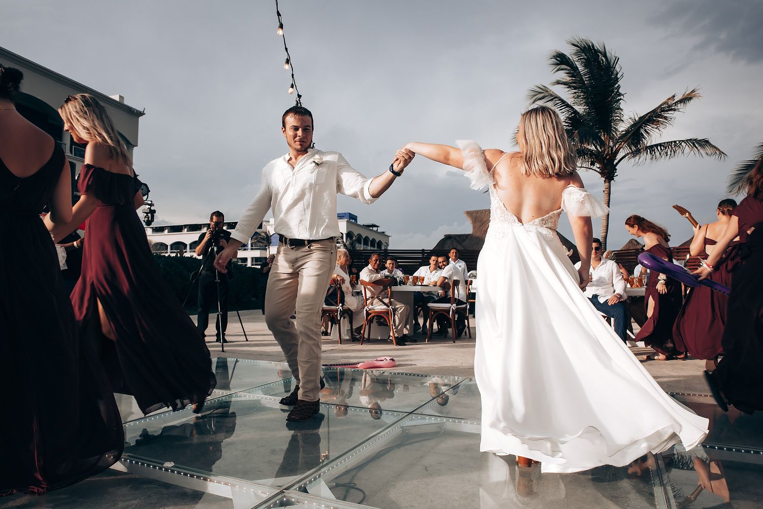 Wedding couple dancing on glass platform with palm trees and ocean backdrop.