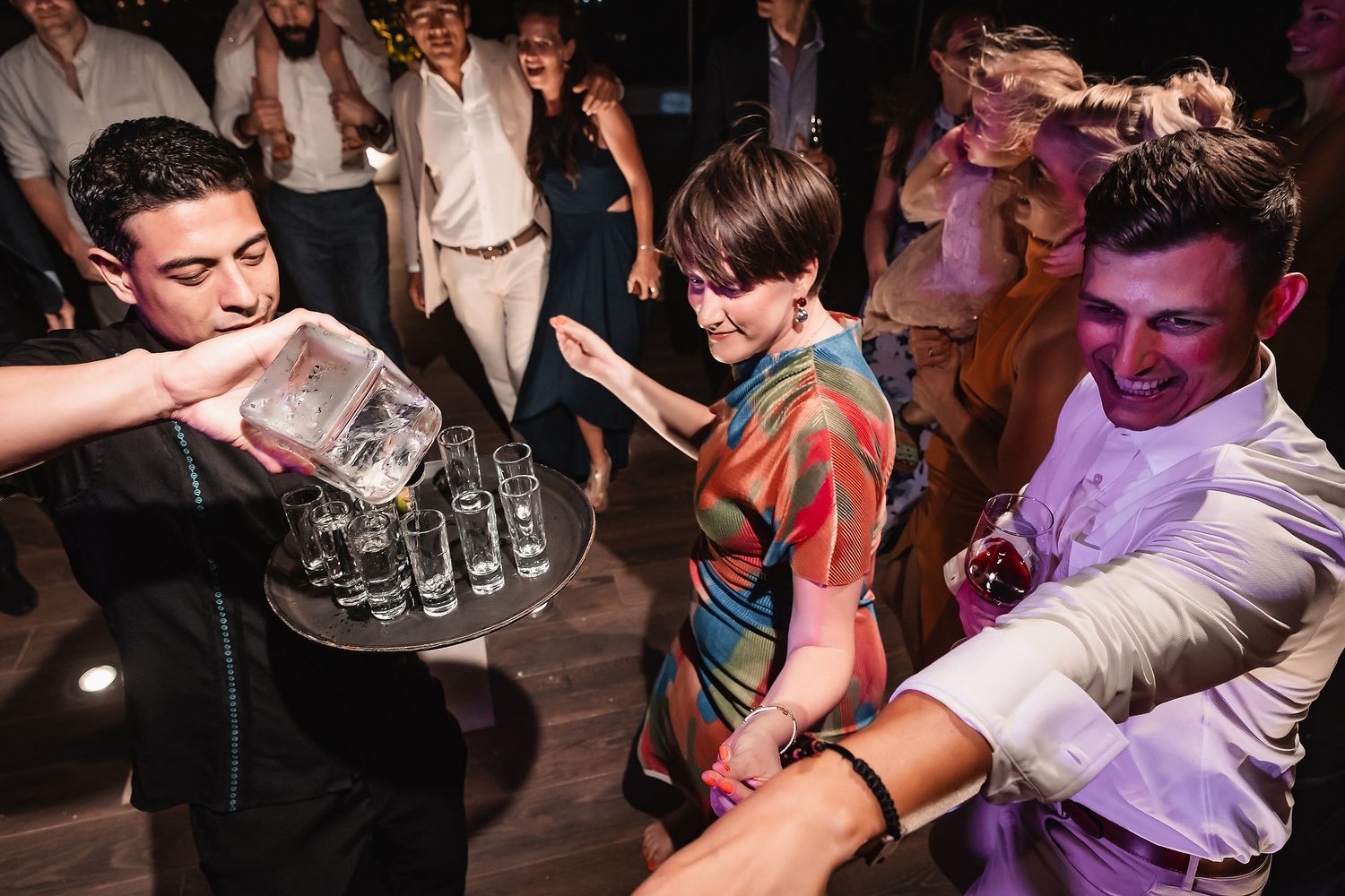 Party guests pour drinks and dance energetically during wedding reception celebrations.