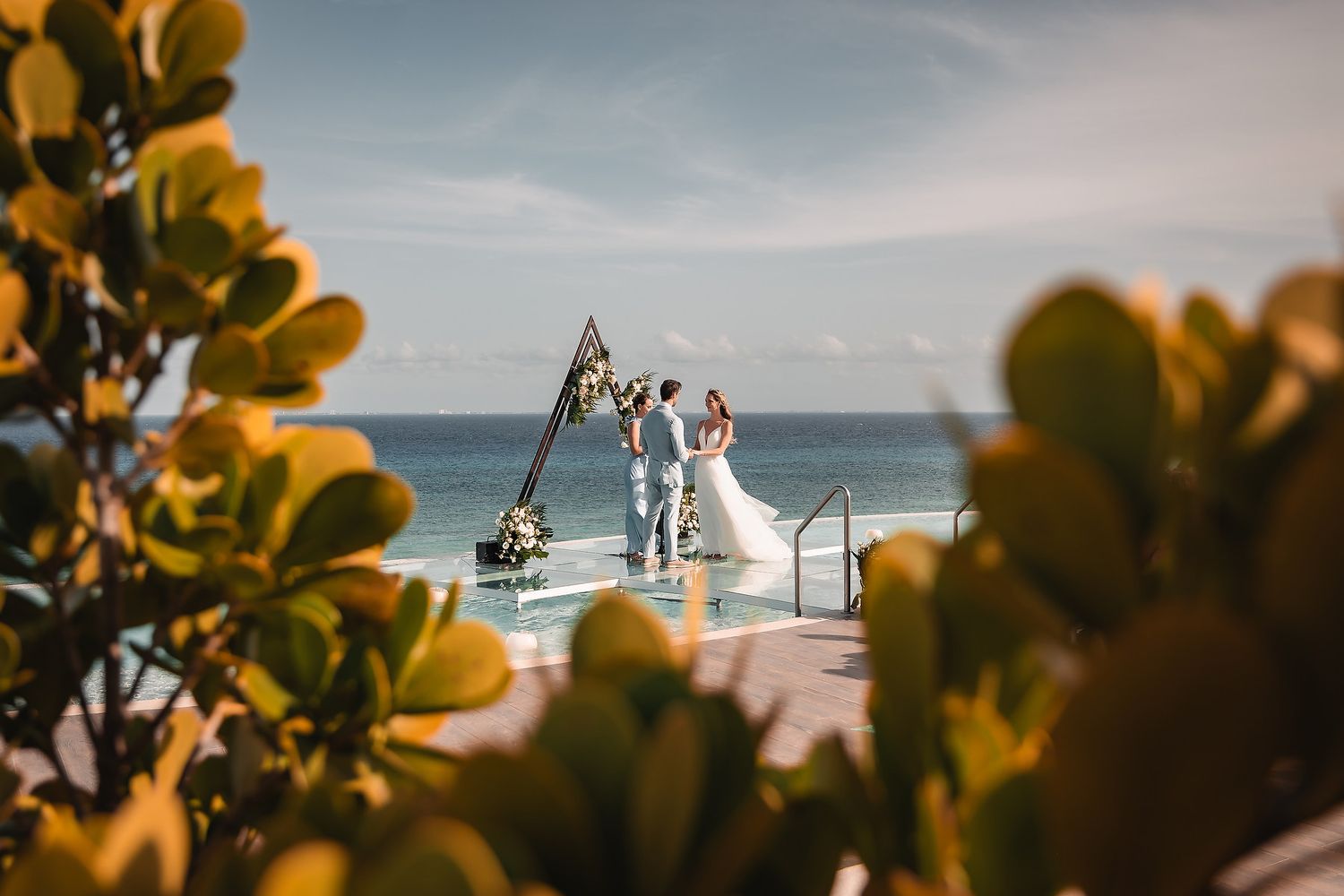 Wedding ceremony viewed through golden leaves with geometric arch and ocean backdrop.