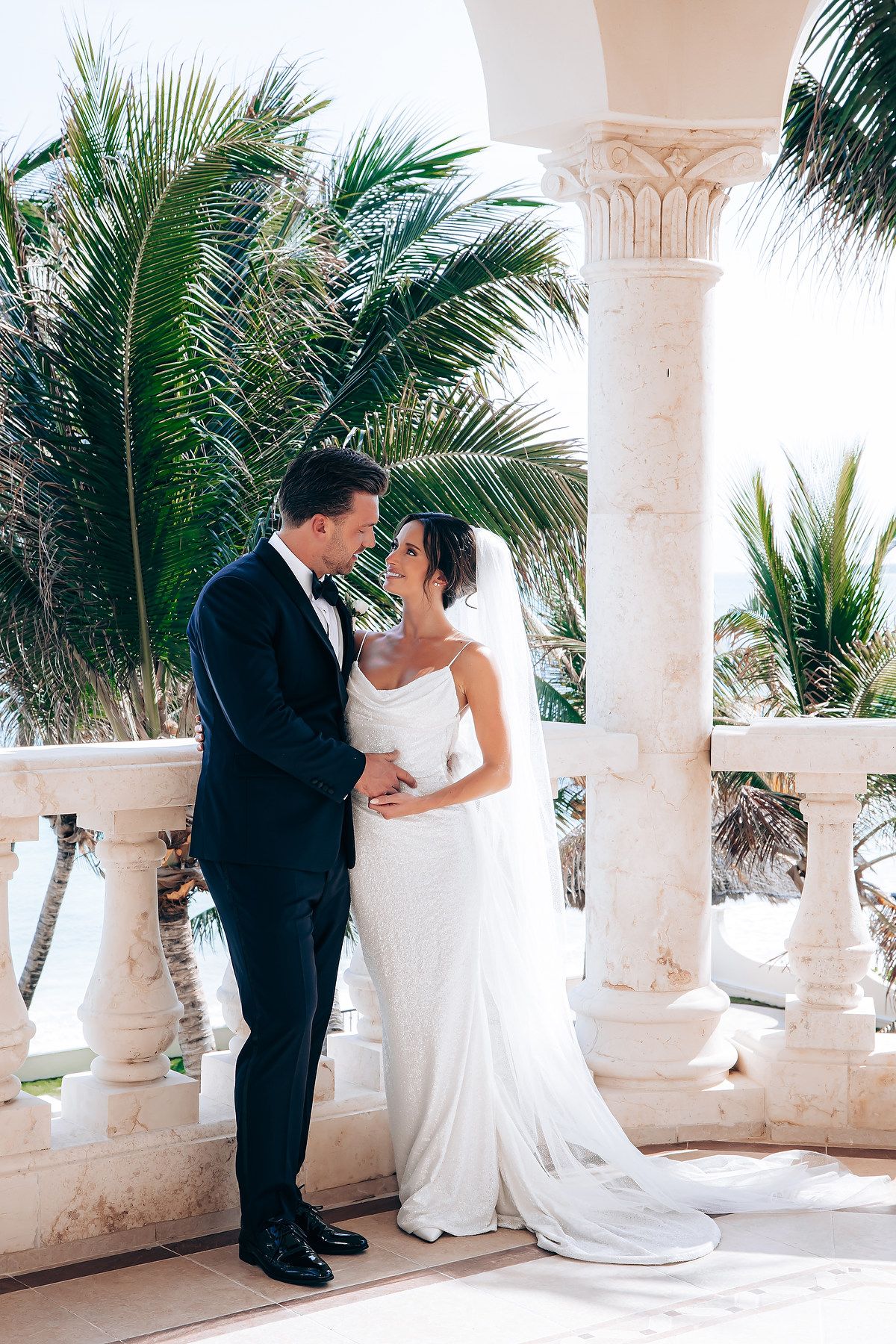 Wedding couple poses along a tropical colonnade with white pillars and palm trees.