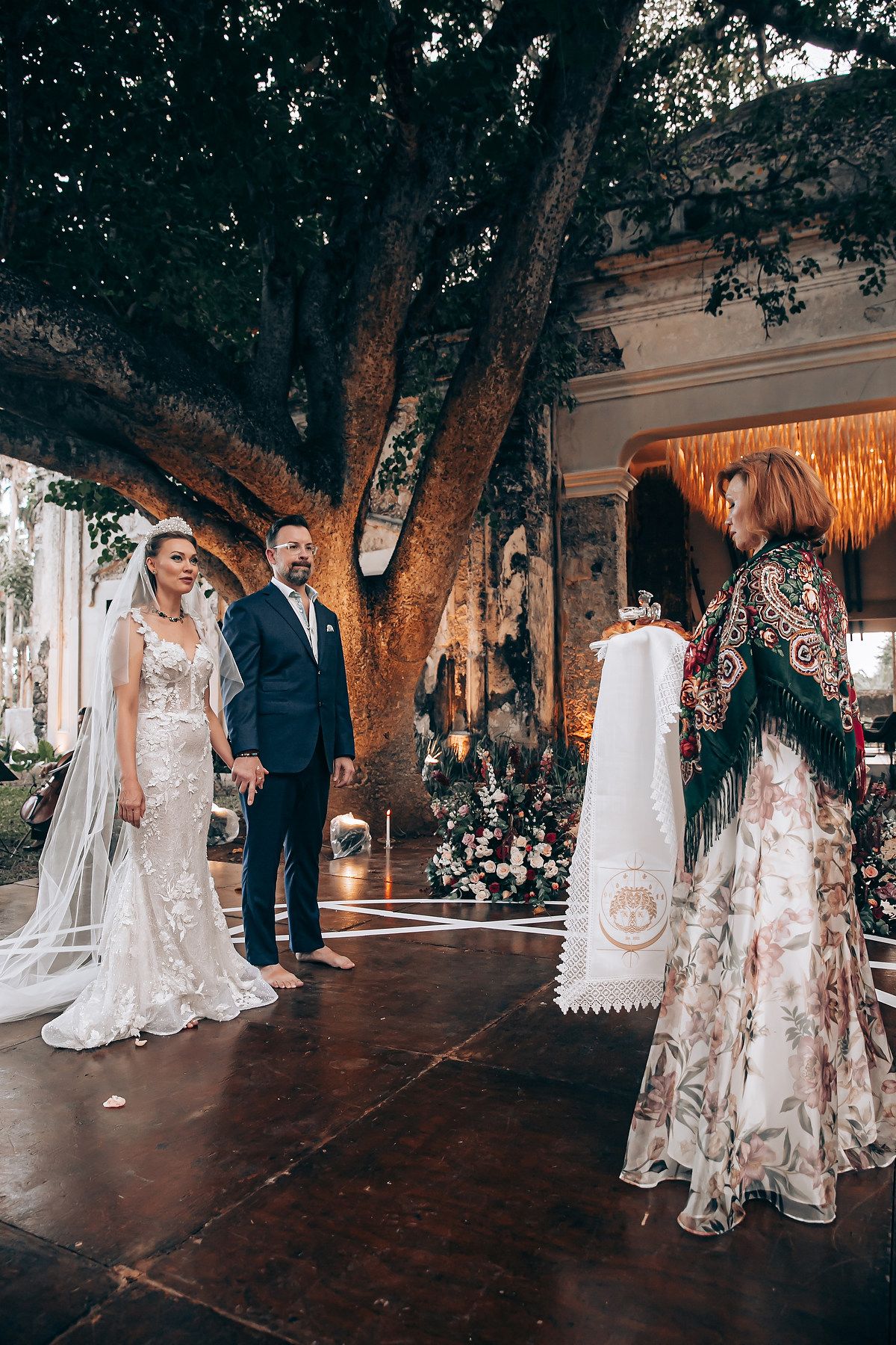 Elegant wedding party poses under trees on a dark wooden deck with romantic lighting.