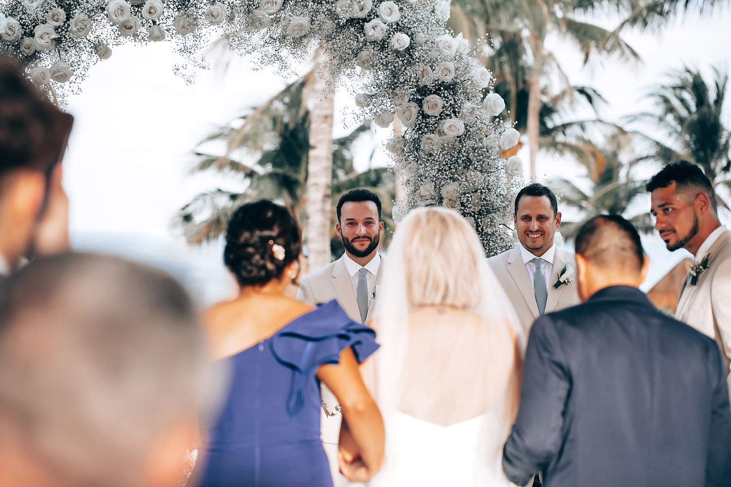 Wedding ceremony under a floral arch with palm trees and tropical decor in an outdoor setting.