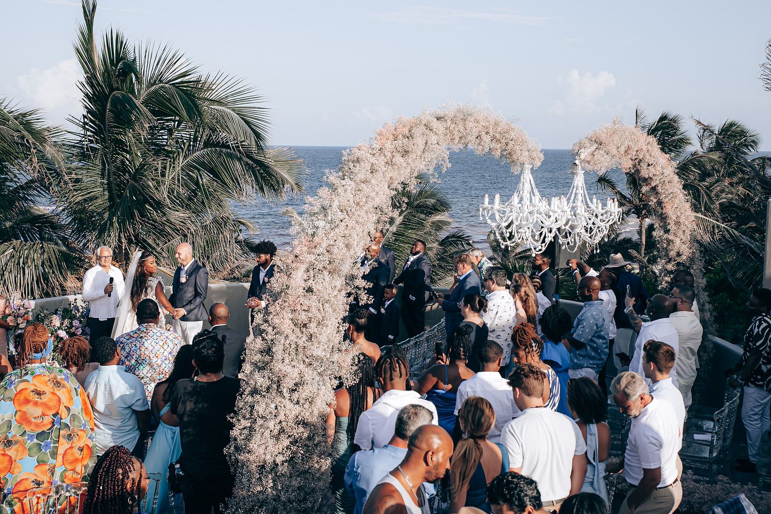A wedding celebration under a floral arch with palm trees and ocean views as guests gather around.