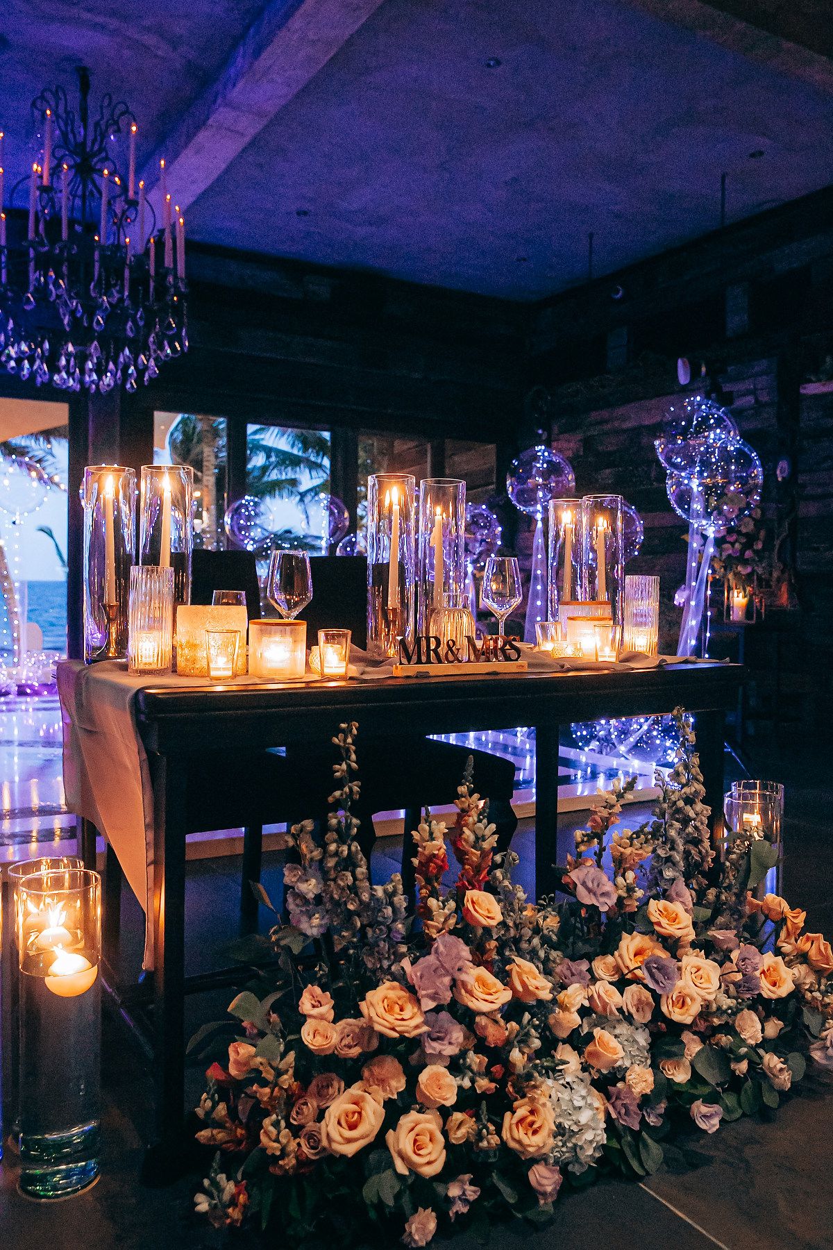 Elegant wedding reception decor with candlelight, crystal chandeliers, and lush floral arrangements in moody purple lighting.