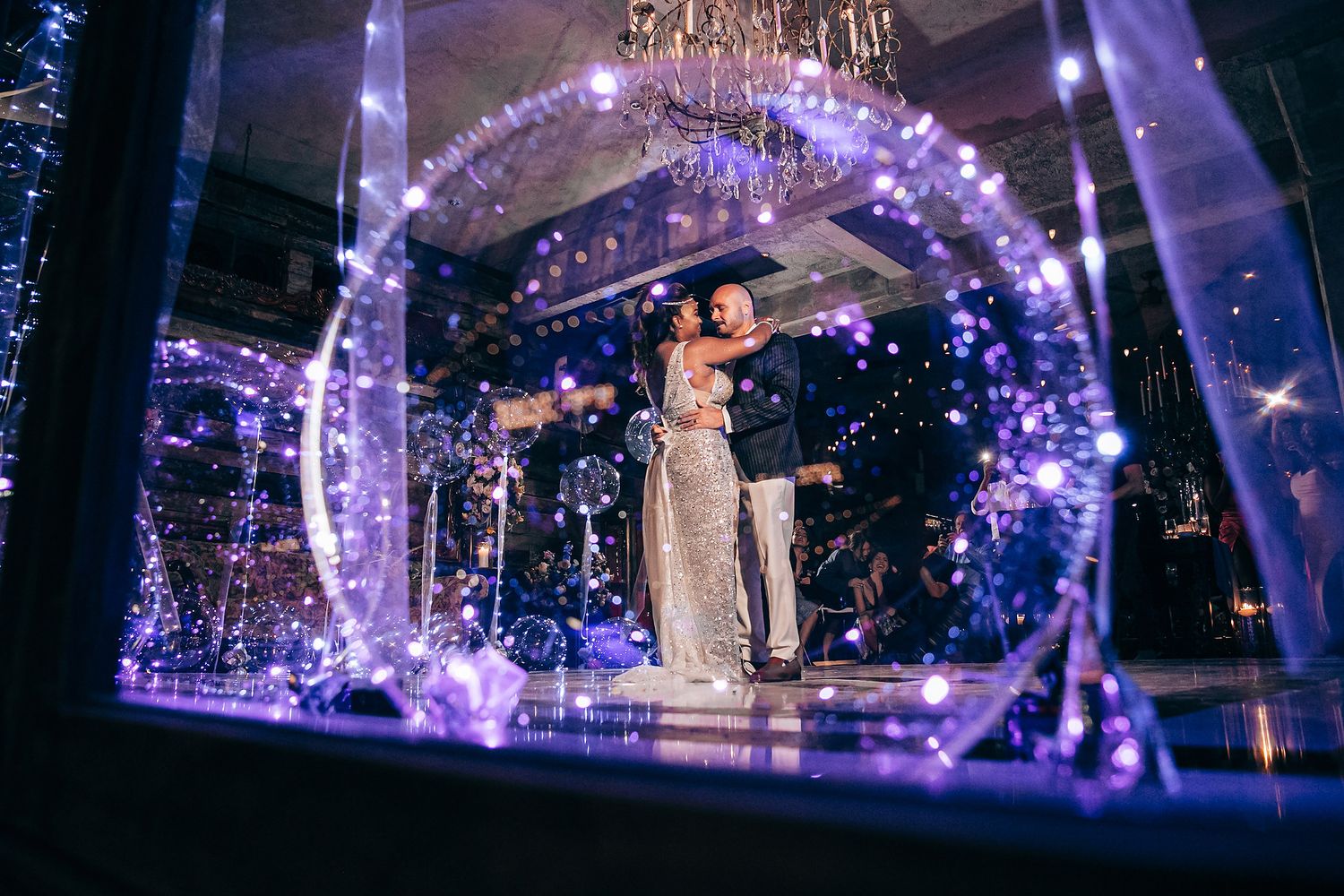 Couple dancing under a glowing purple archway with sparkling lights and a chandelier in an elegant venue.