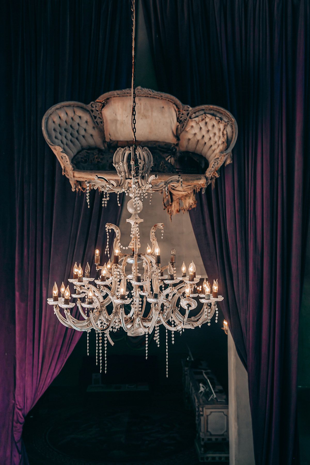 An elegant crystal chandelier hangs beneath a tufted vintage sofa against dark velvet curtains in a theatrical setting.