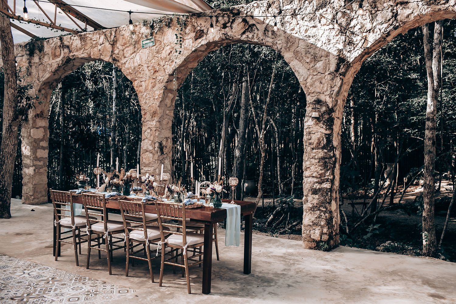 Elegant dining table set beneath weathered stone arches with forest backdrop creates rustic ambiance.