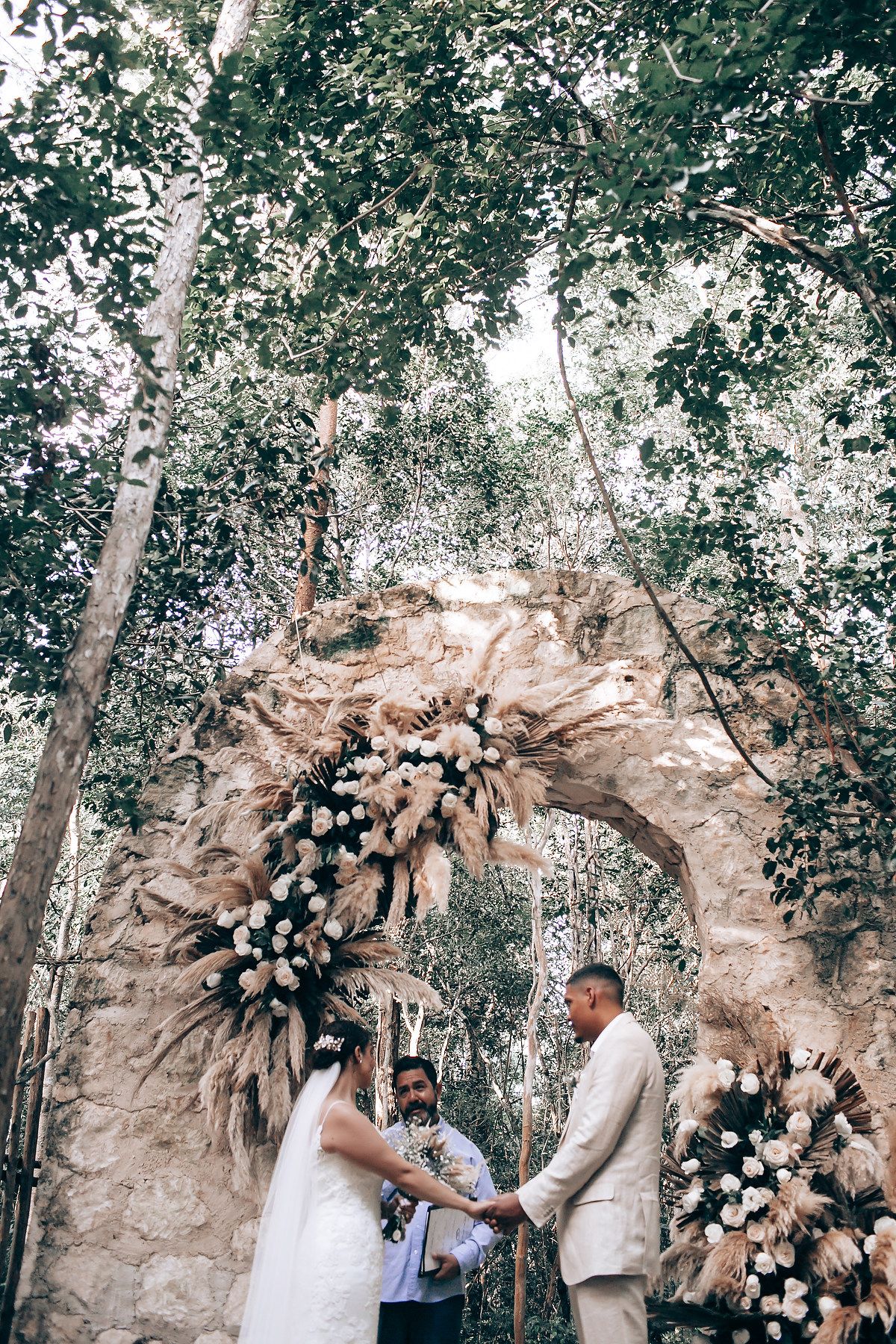 A romantic wedding ceremony takes place under a floral arch in a wooded outdoor setting with natural lighting.