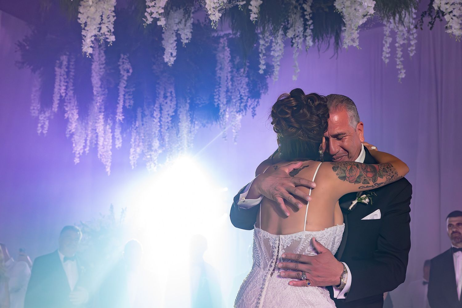 Newlyweds dancing together under cascading white floral arrangements with dramatic purple stage lighting.