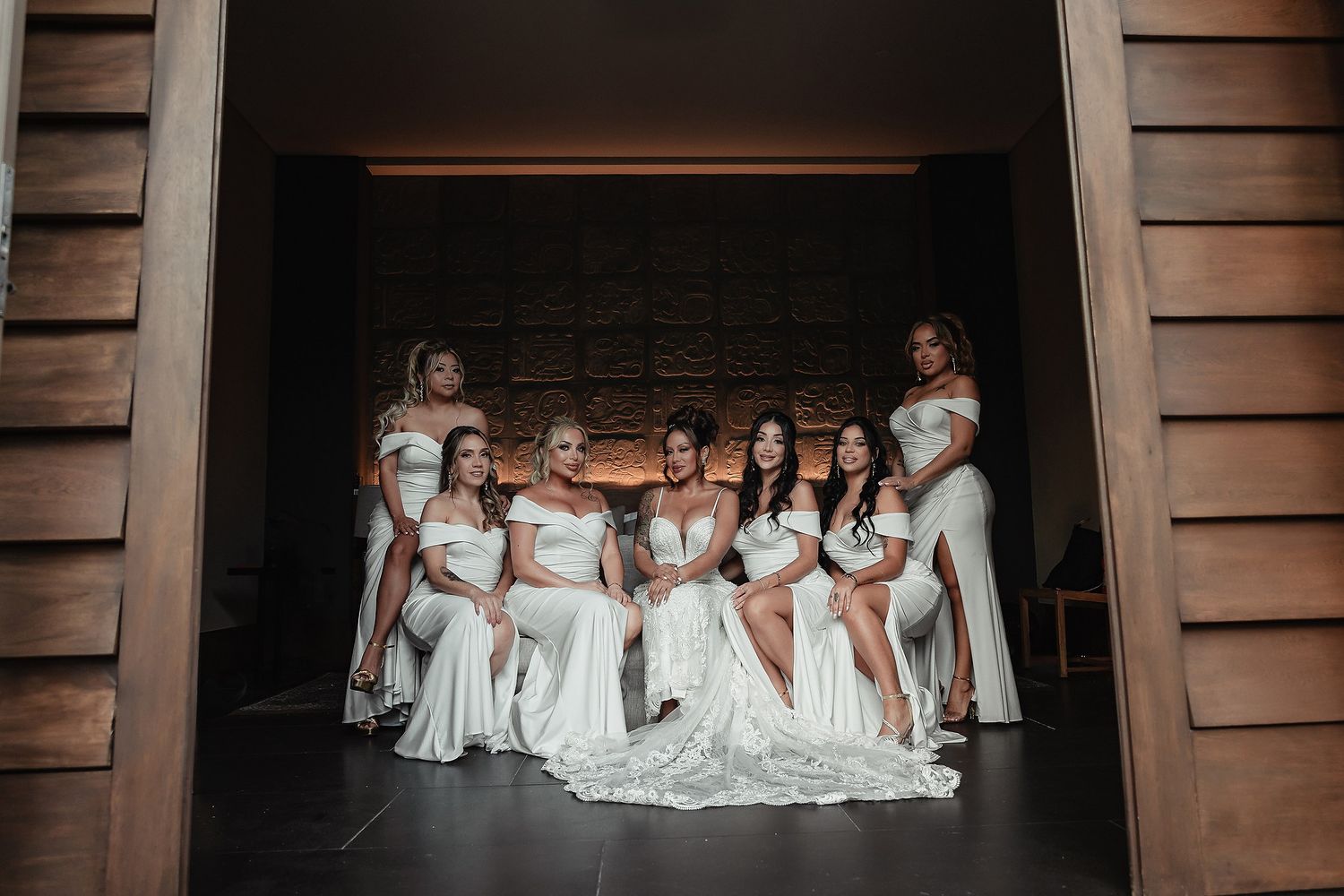Group wearing white dresses sitting together on wooden floor in dramatic lighting.