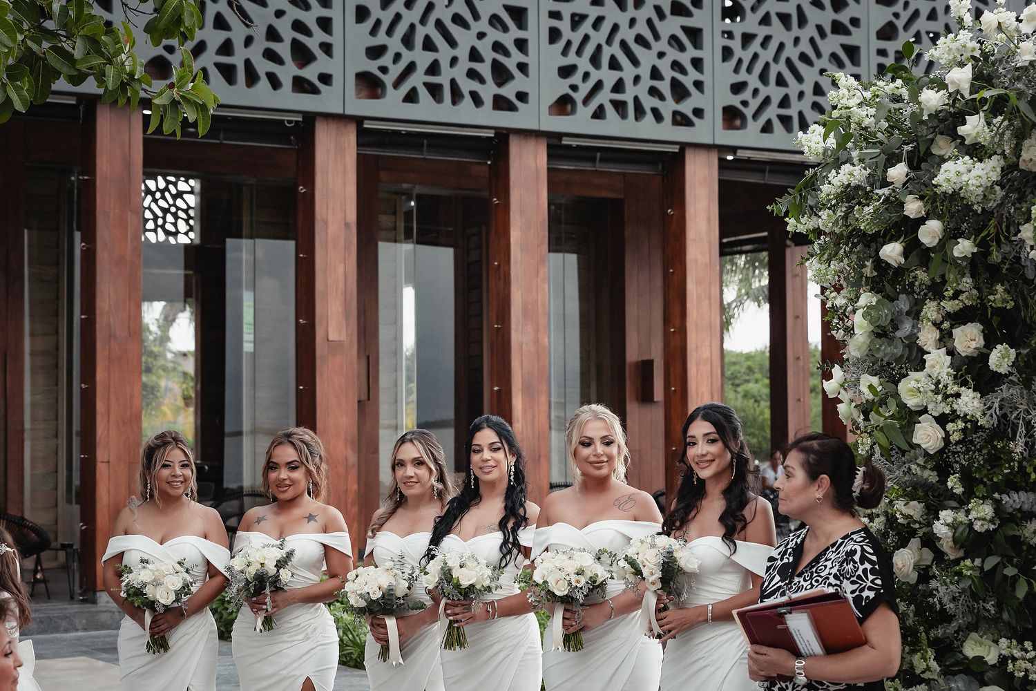Wedding party in formal attire stands in a row against an ornate architectural backdrop with white floral arrangements.