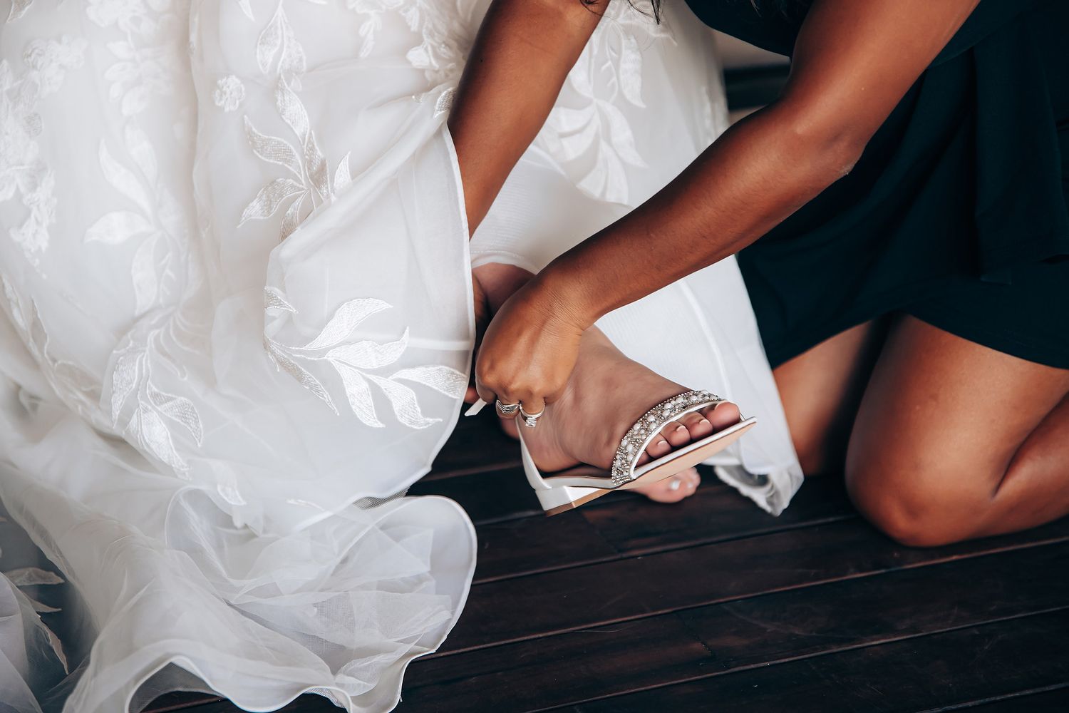 Hands adjusting a flowing white wedding dress train on a dark wooden floor.