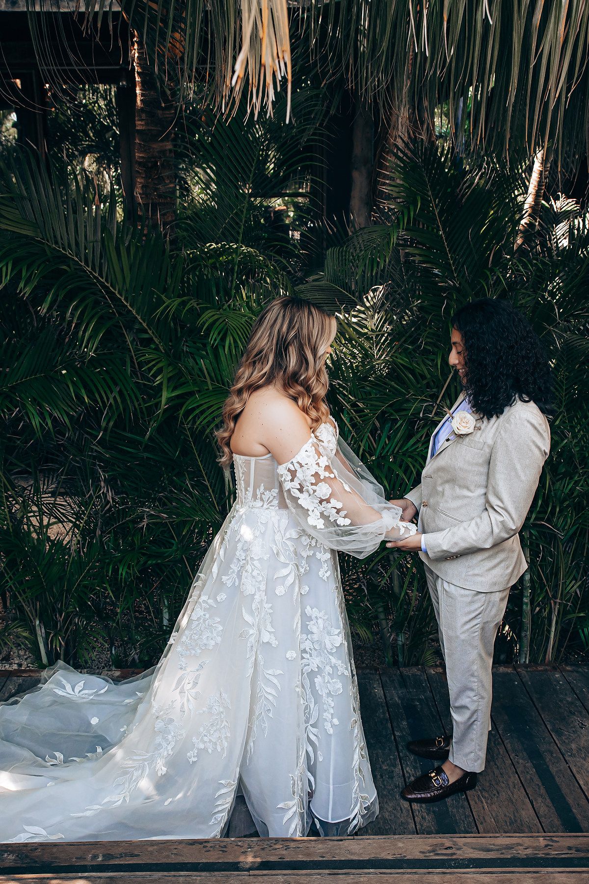 A romantic wedding photoshoot on wooden steps with a flowing white dress and gray suit among tropical greenery.
