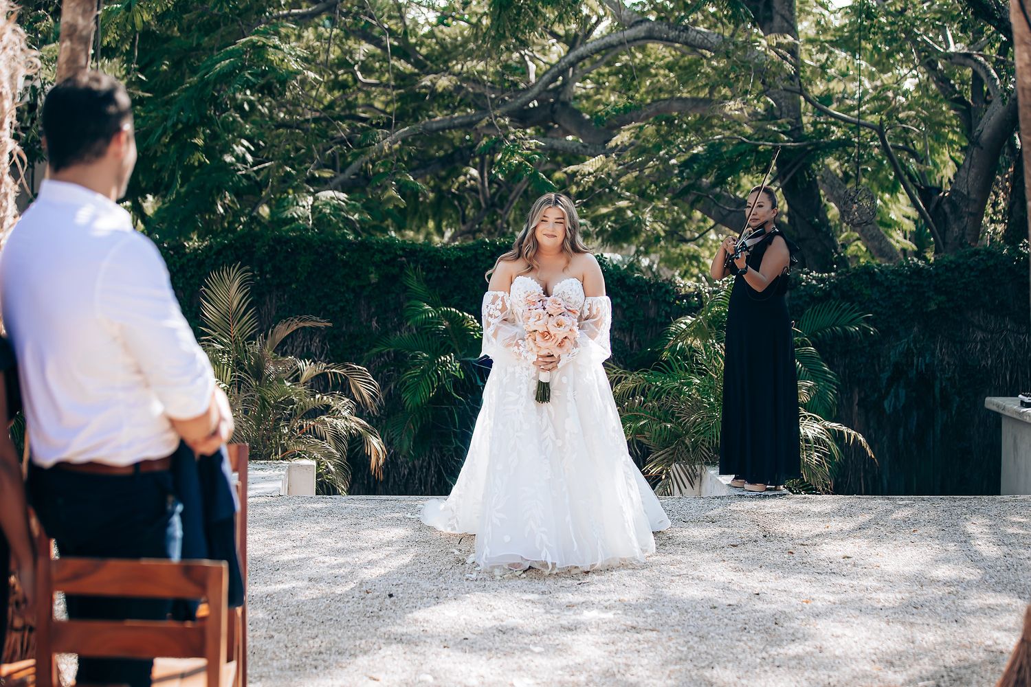 Wedding ceremony guests gather outdoors under tall trees on a sunny day.