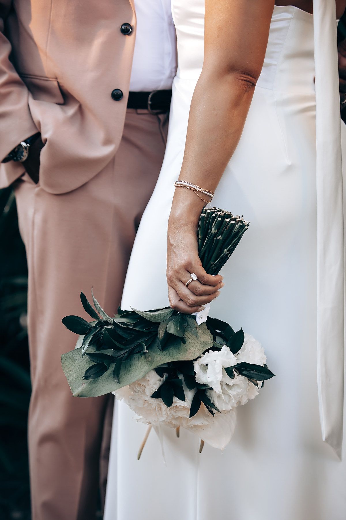 Close-up of wedding attire showing a blush pink suit and white dress with a black and white bouquet.