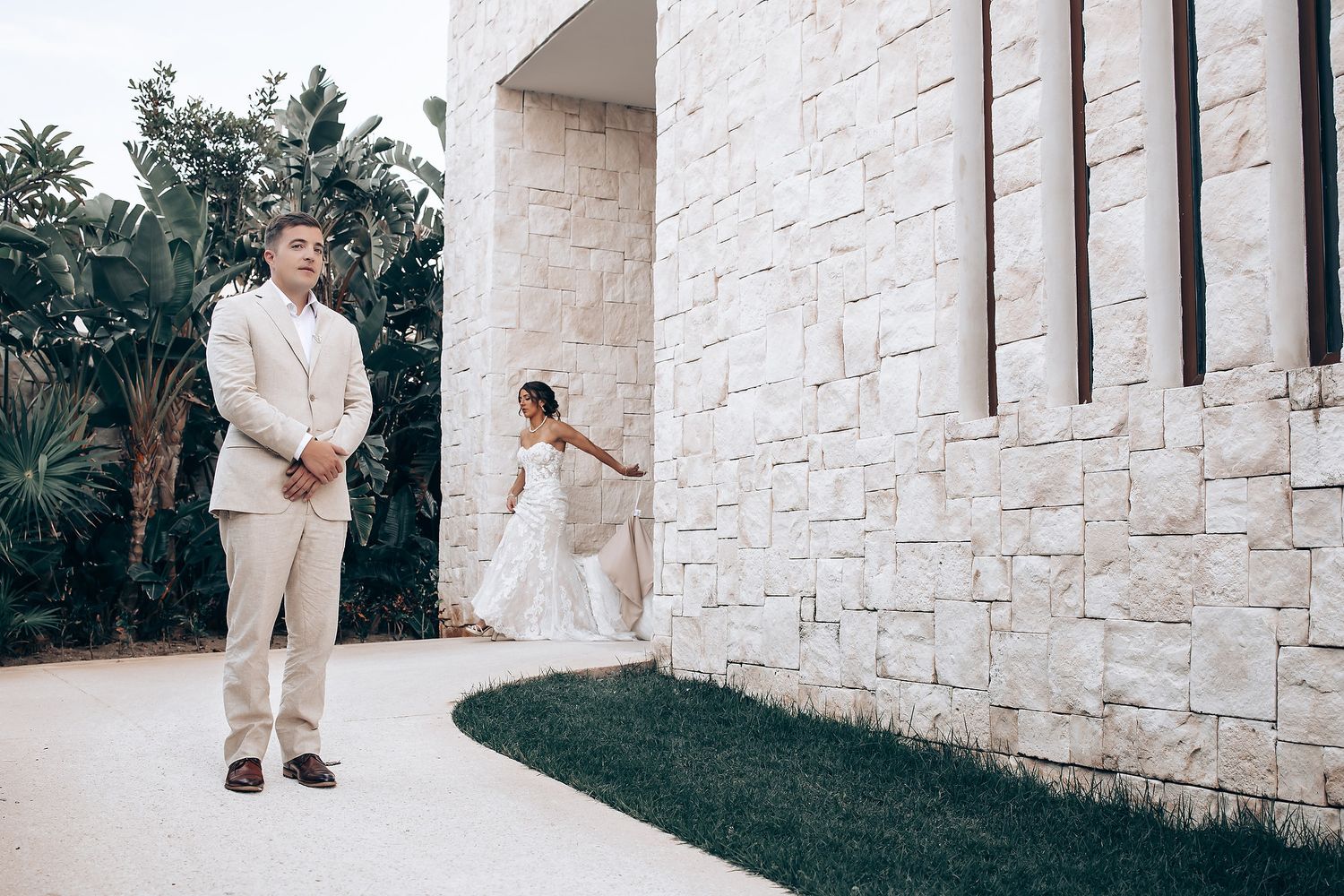 Couple in light colored formal attire poses against stone wall exterior of building.