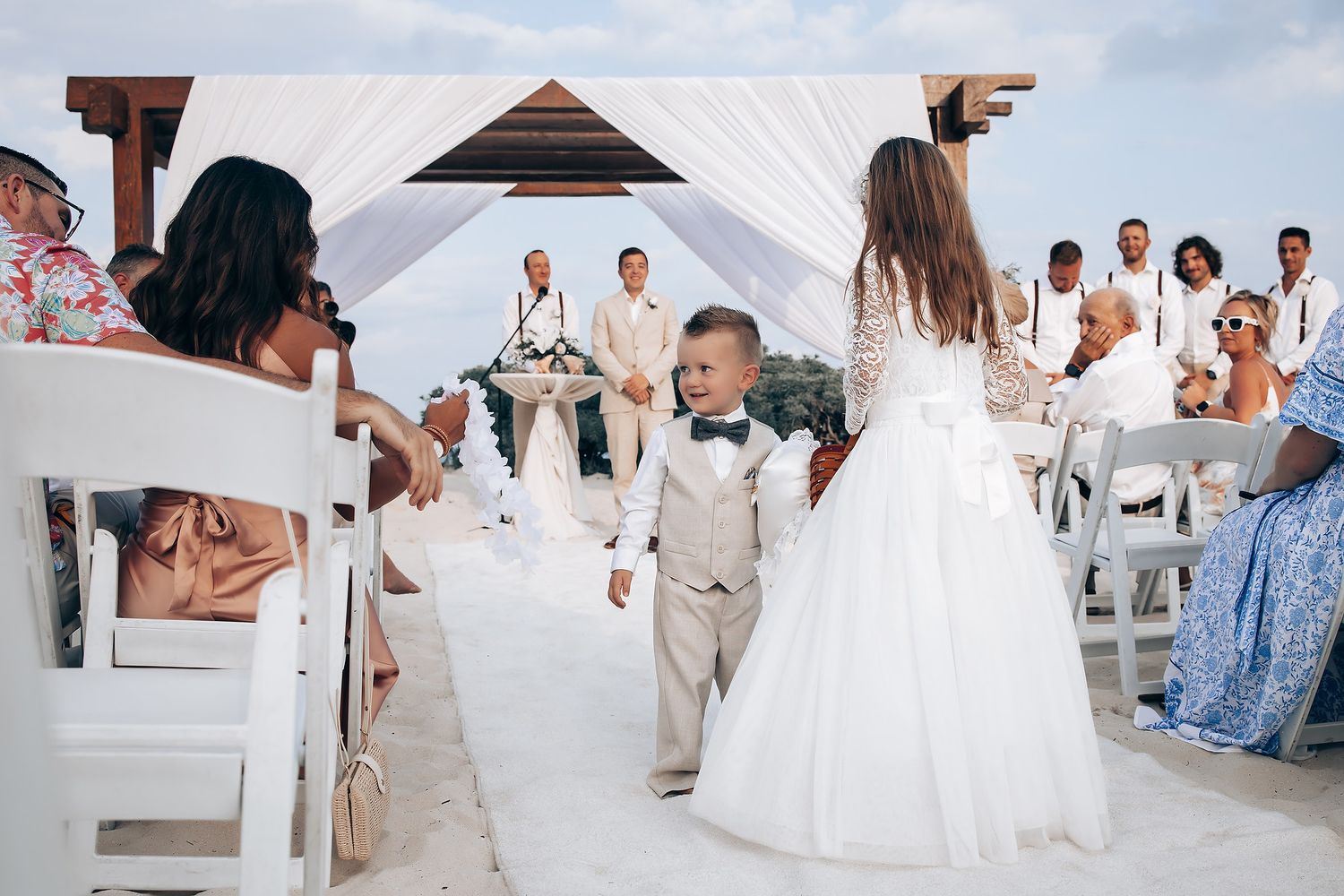 Wedding guests celebrate and dance at a beachside reception with white decor and tropical backdrop.