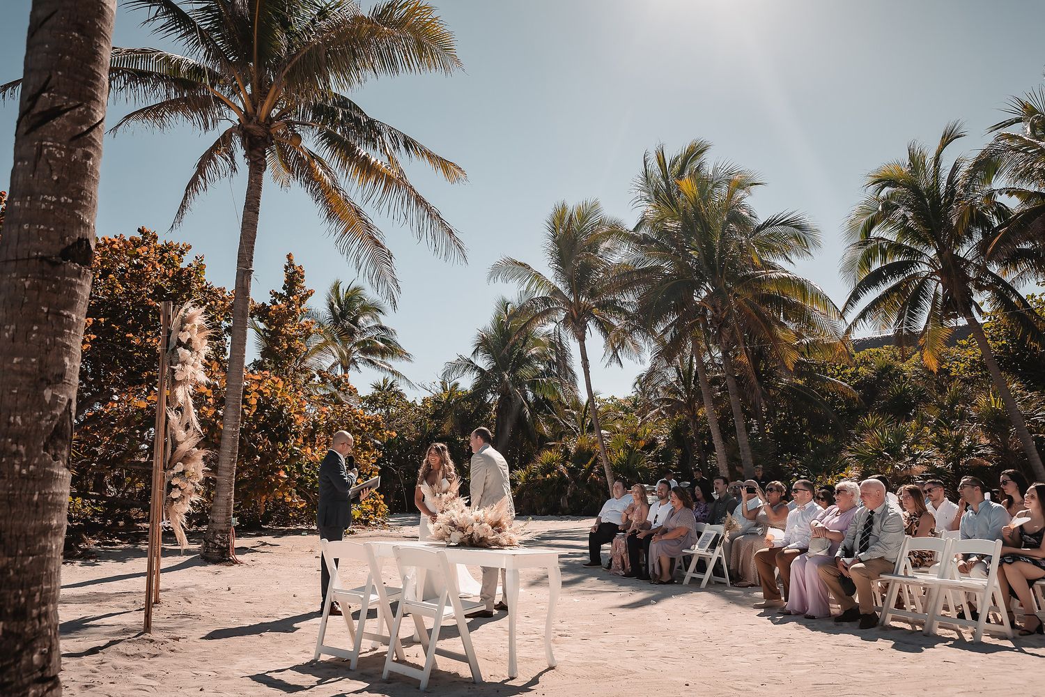 A scenic beach wedding ceremony setup with palm trees and white chairs arranged on a sandy path.