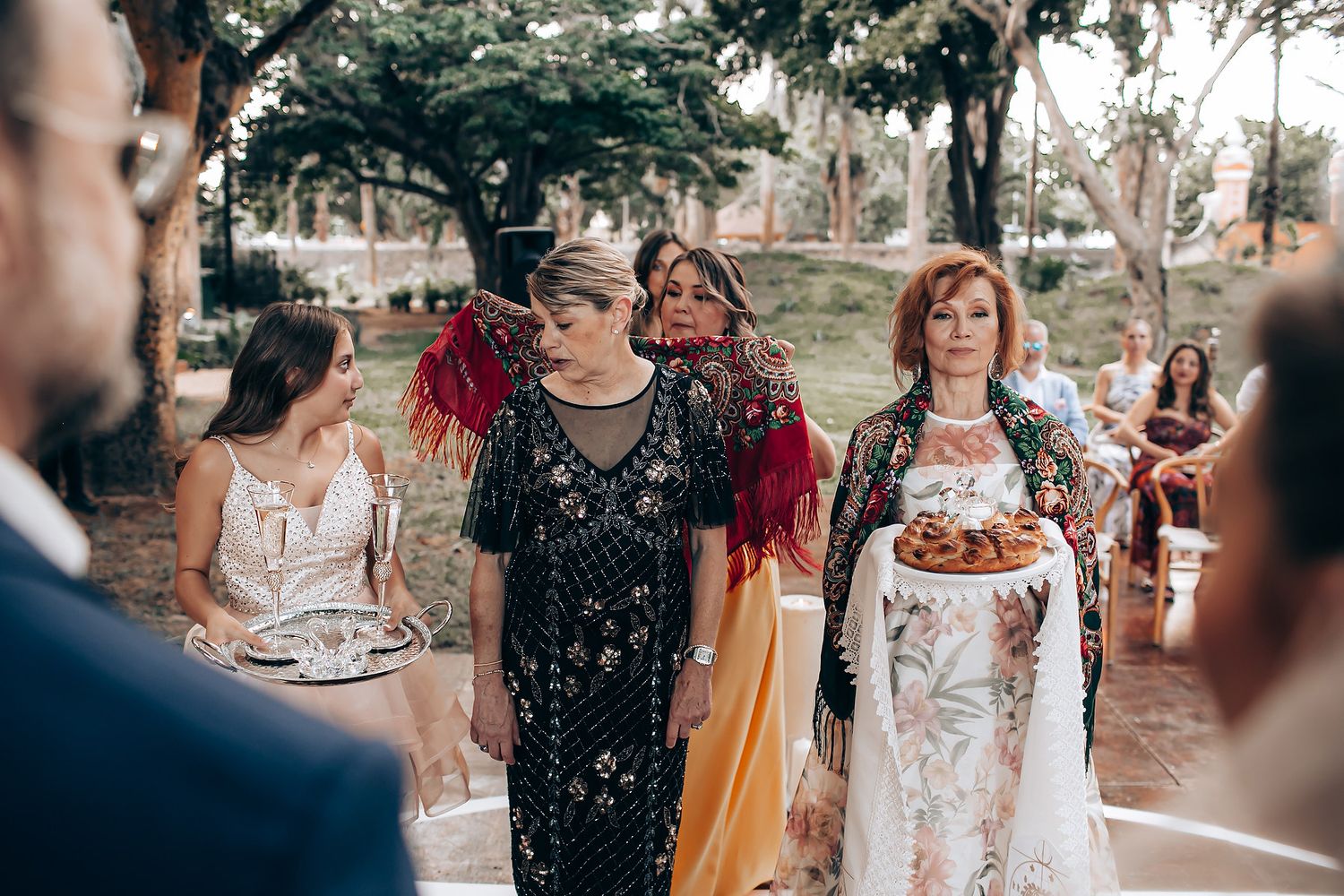 A group of wedding guests gathered outdoors at a rustic venue with trees and string lights in the background.