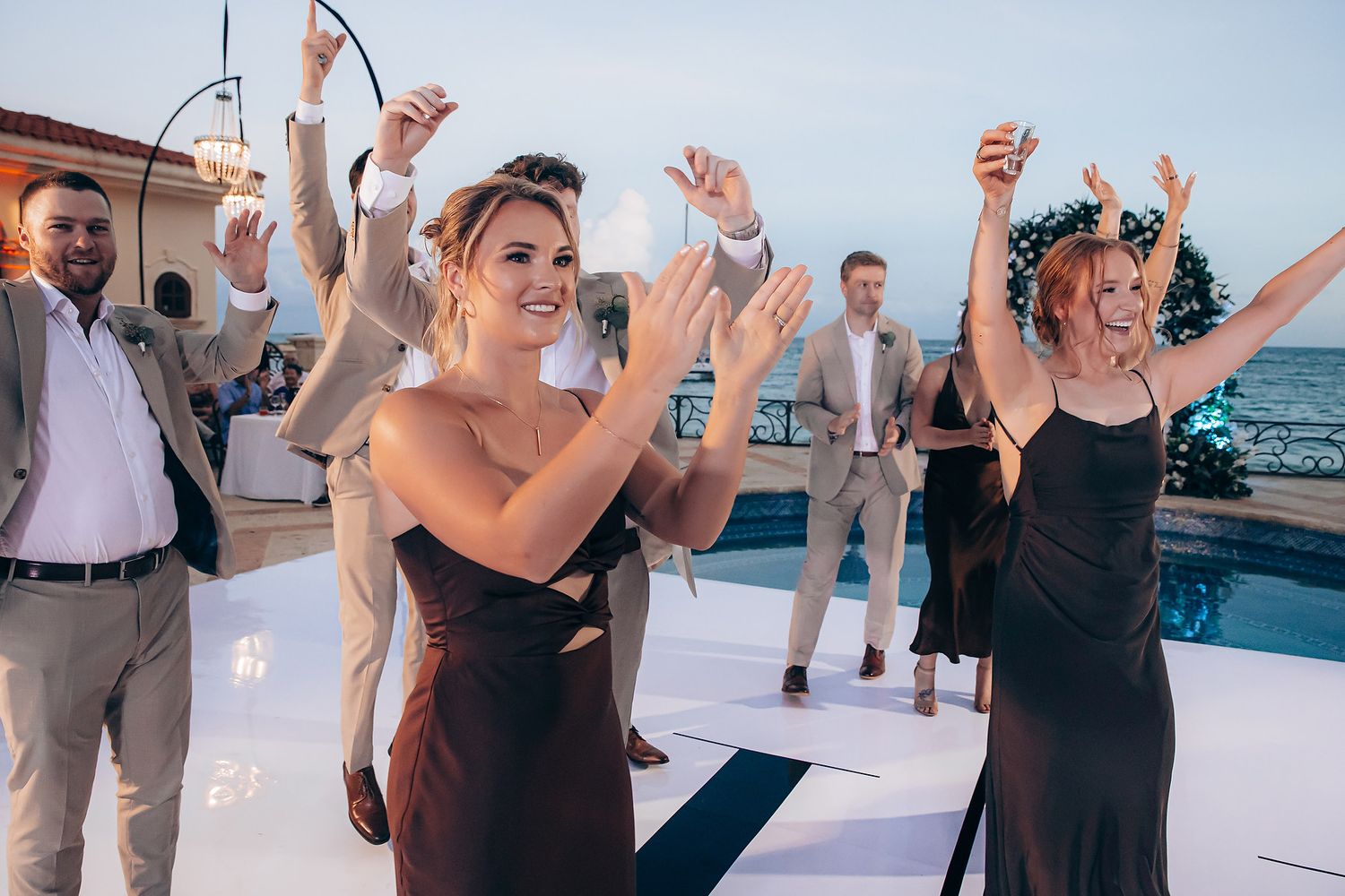 Wedding guests dancing and celebrating on a white dance floor at an outdoor beachside reception.