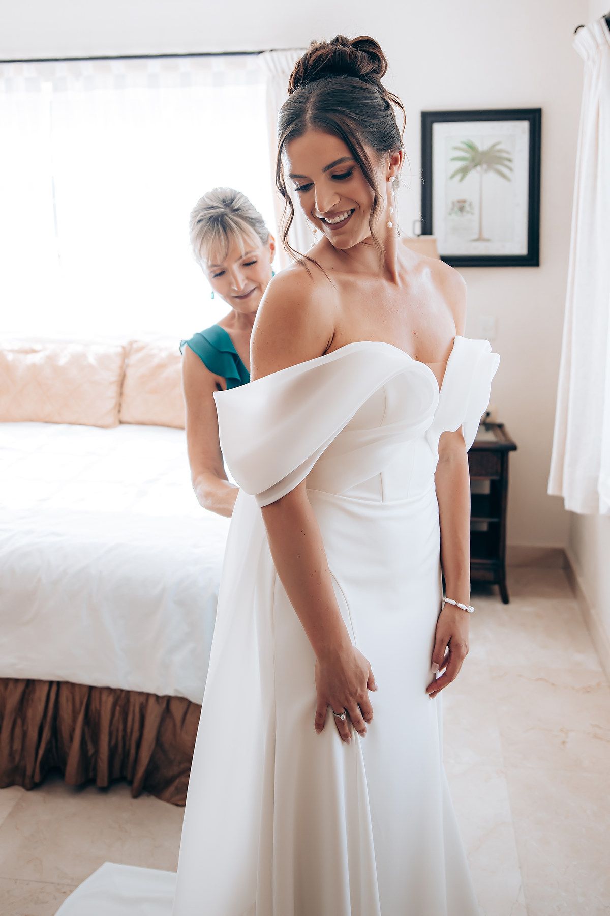 Two women in elegant white dresses smiling and adjusting wedding gown in bright bedroom.