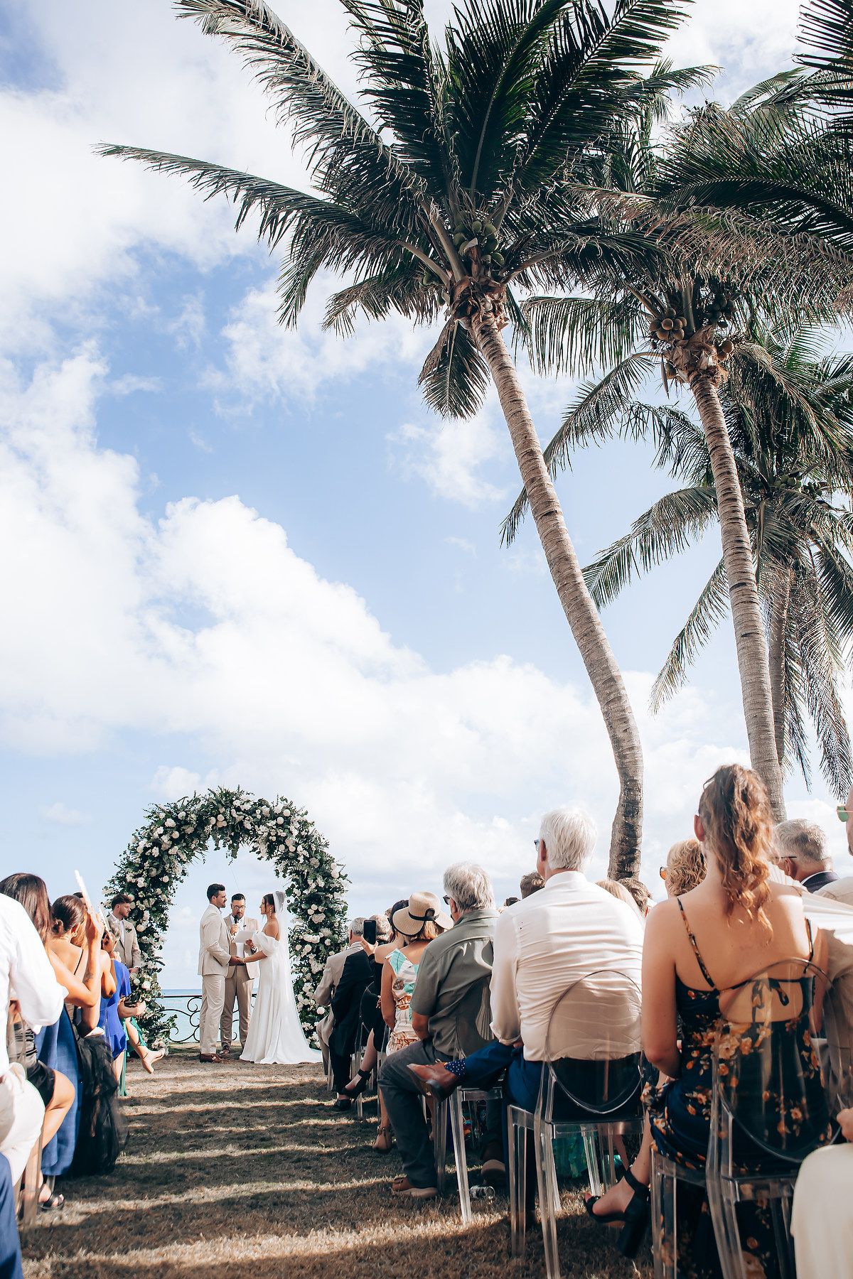 Wedding guests gather under swaying palm trees at a tropical outdoor ceremony on a hilltop overlooking the ocean.