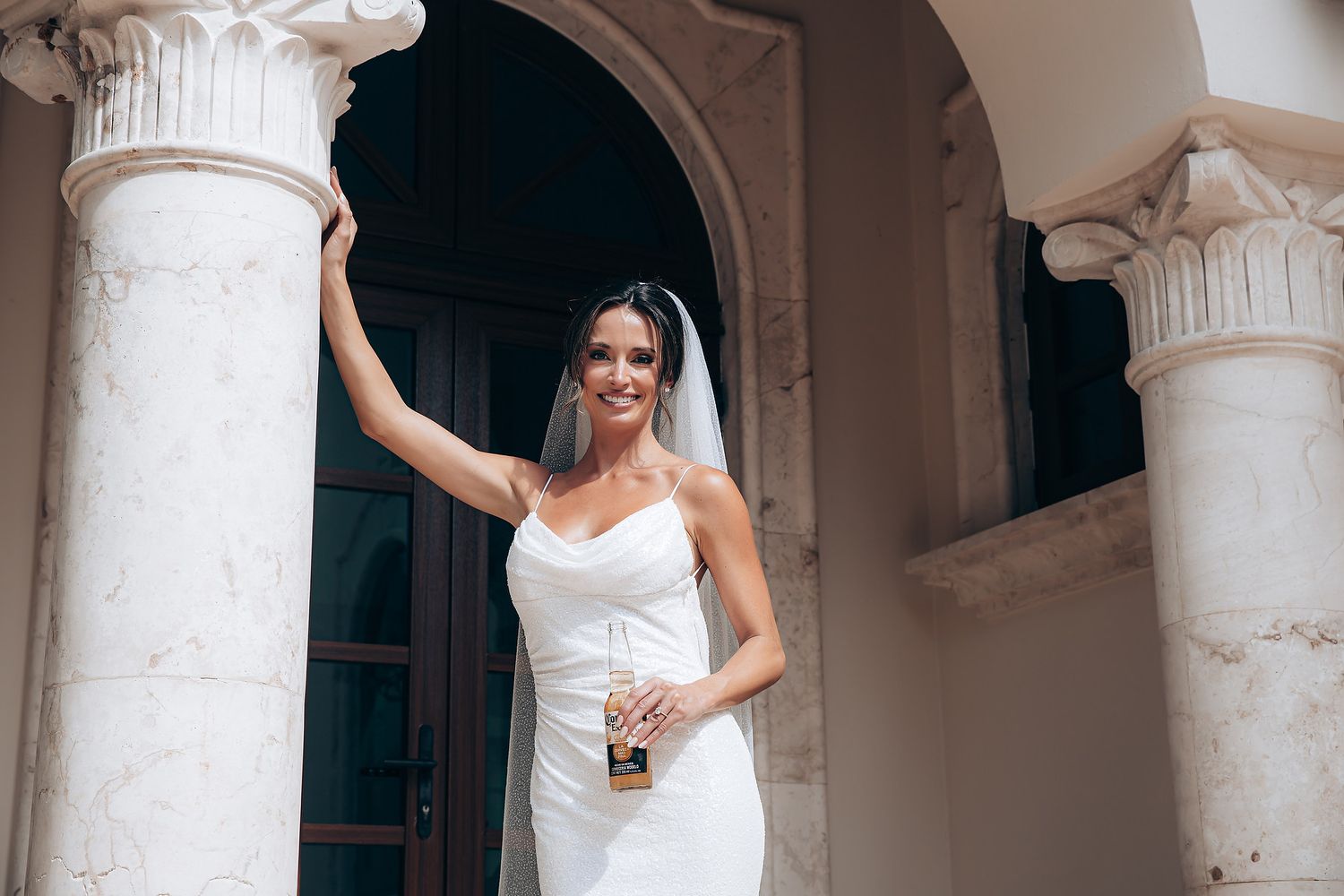 A bride in a white wedding dress poses elegantly between classical white stone columns in an ornate venue.