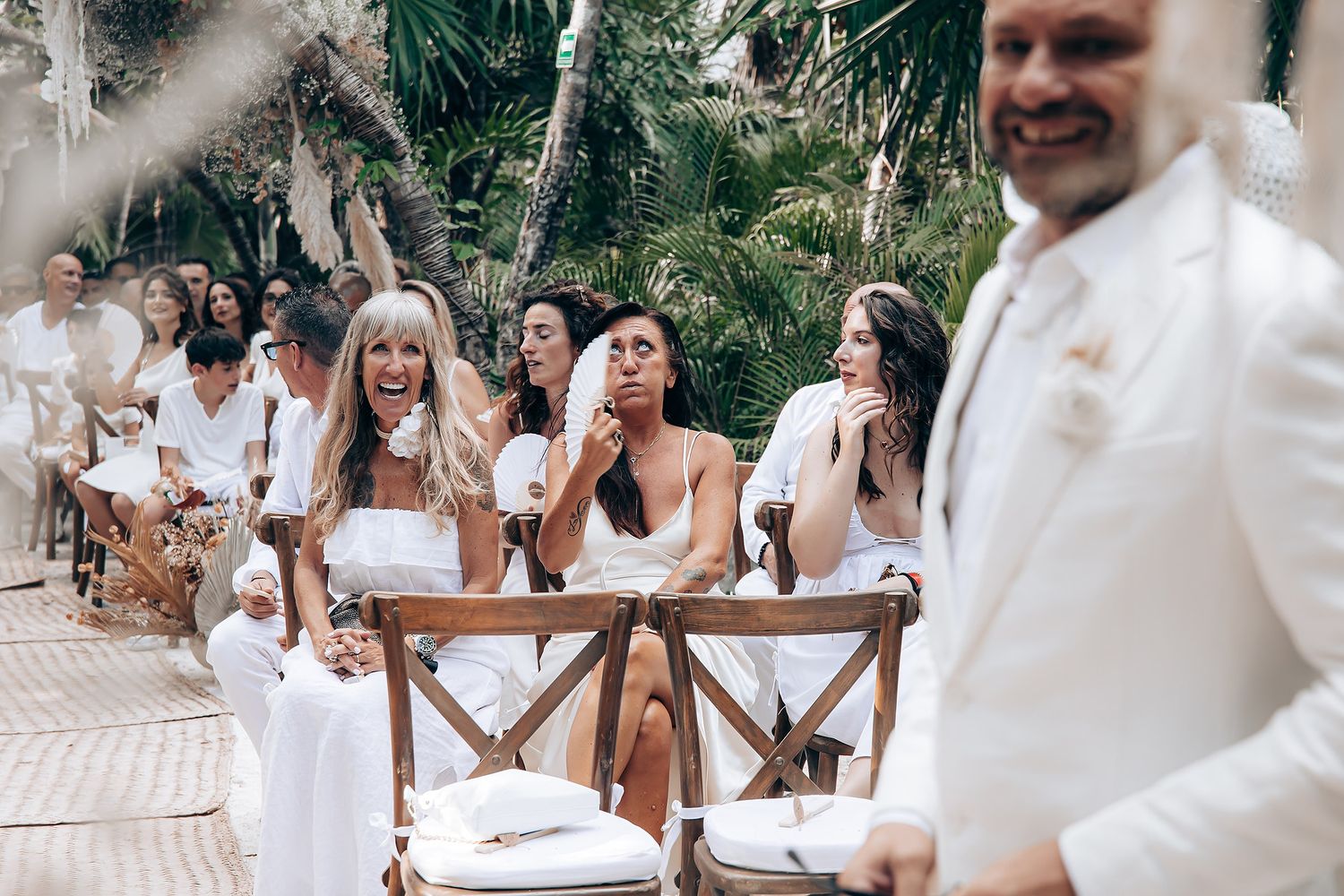 Wedding guests in white attire socialize at an outdoor tropical venue with wooden chairs.