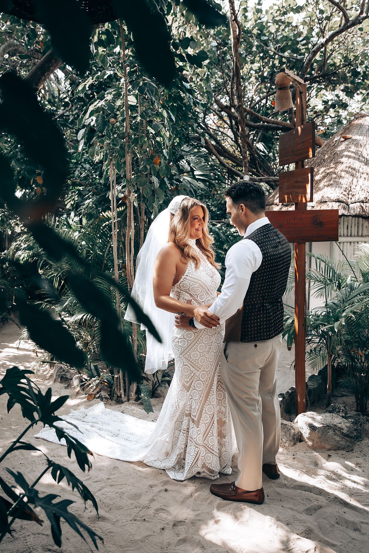 A couple in wedding attire shares an intimate moment together in a tropical garden setting with dappled sunlight.