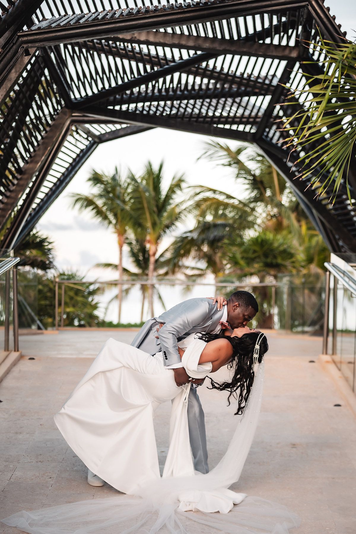 A romantic wedding couple shares a dip kiss under a pergola with palm trees and tropical scenery in the background.