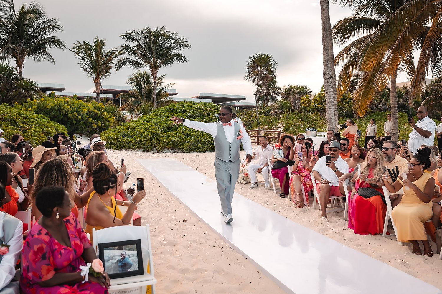 Wedding guests gather on a tropical beach with palm trees and white sand during a sunset ceremony.
