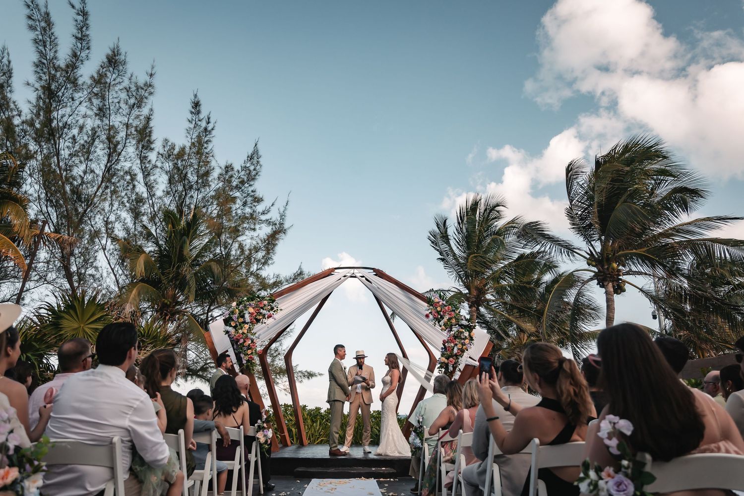 Outdoor beach wedding ceremony with wooden arch and white fabric draped under blue sky and palm trees.