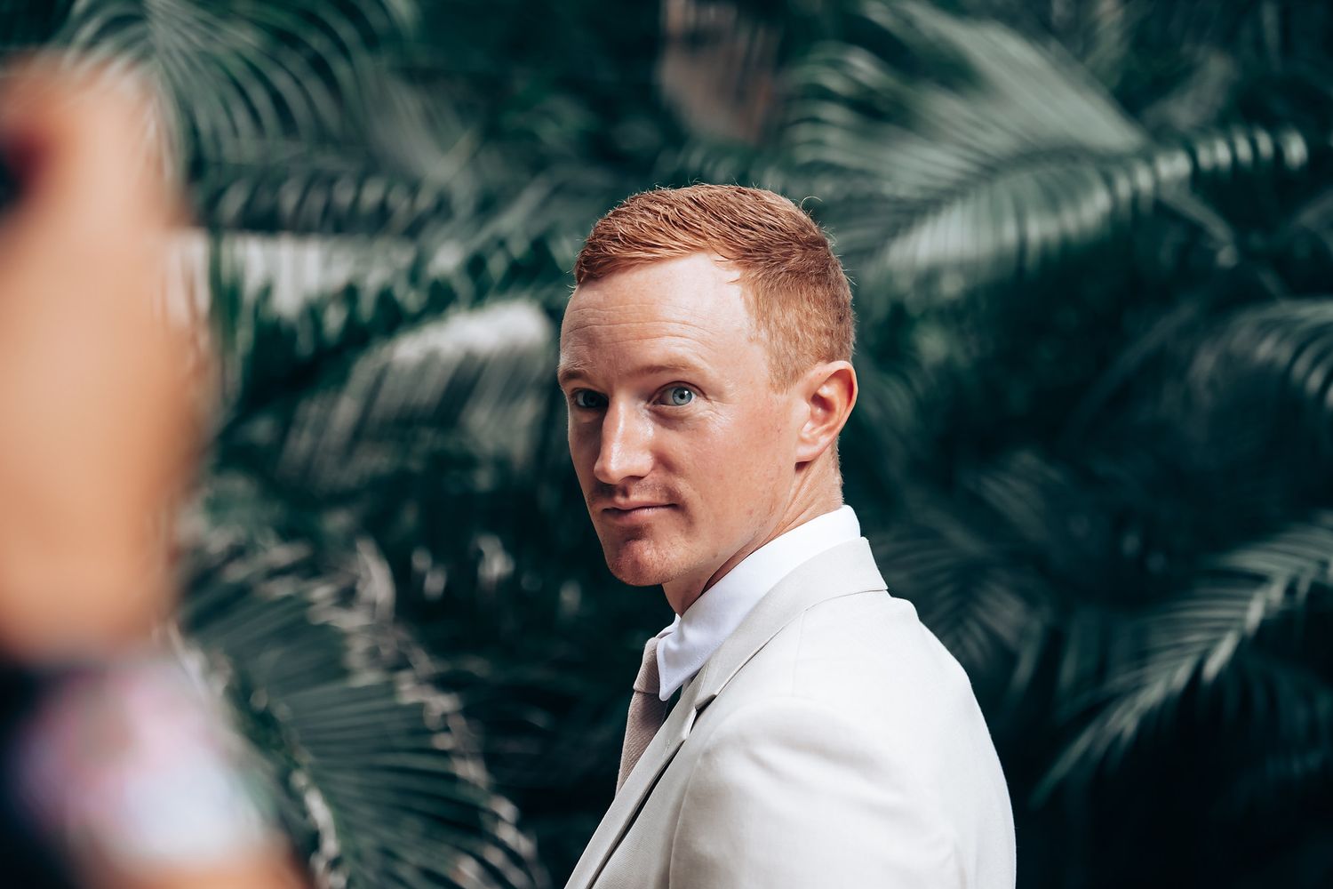A groom in a crisp white suit stands against a backdrop of tropical palm leaves.