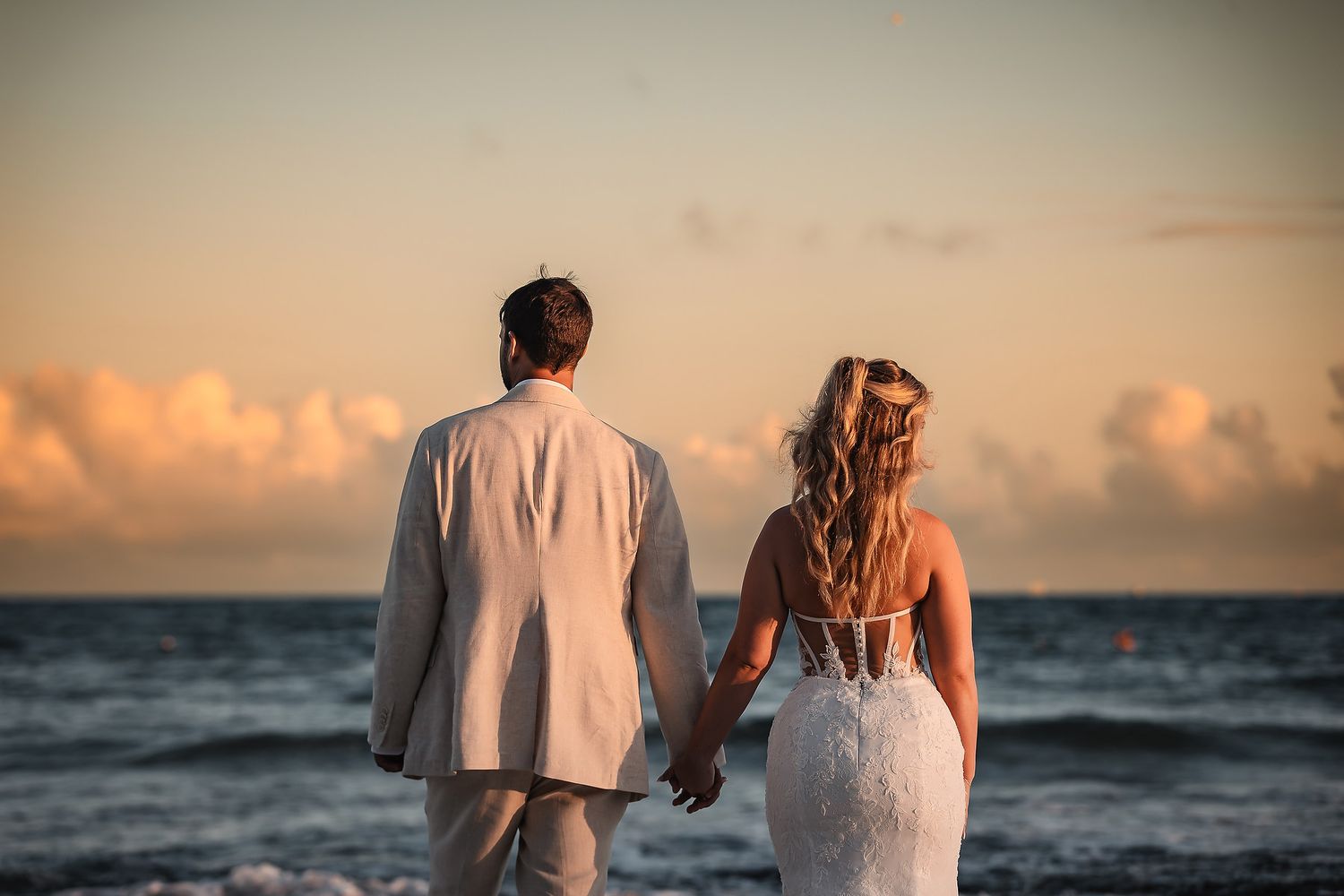 Newlyweds walk along a beach at sunset with ocean waves in the background.