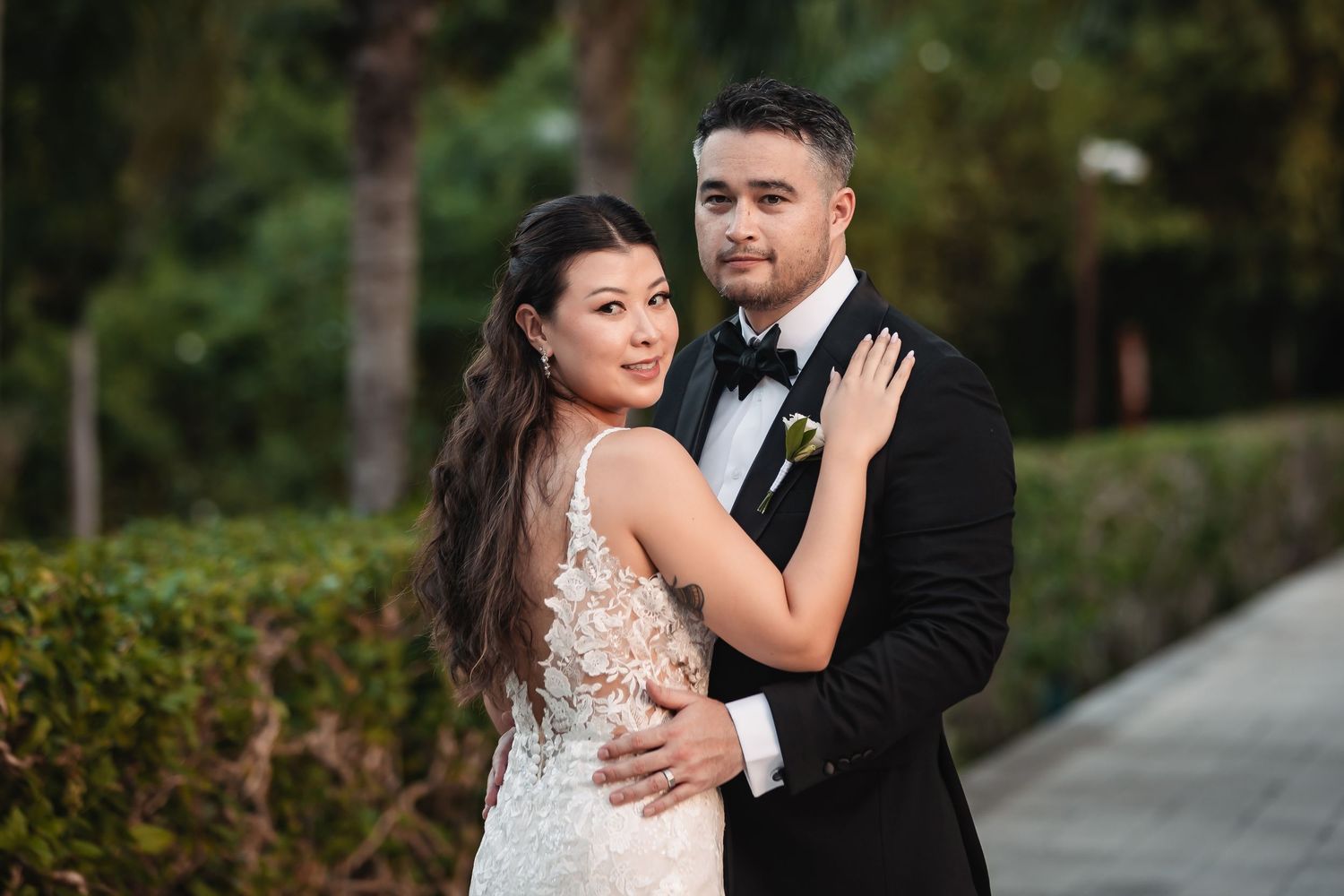 Elegant wedding couple poses in formal attire against tropical garden backdrop.